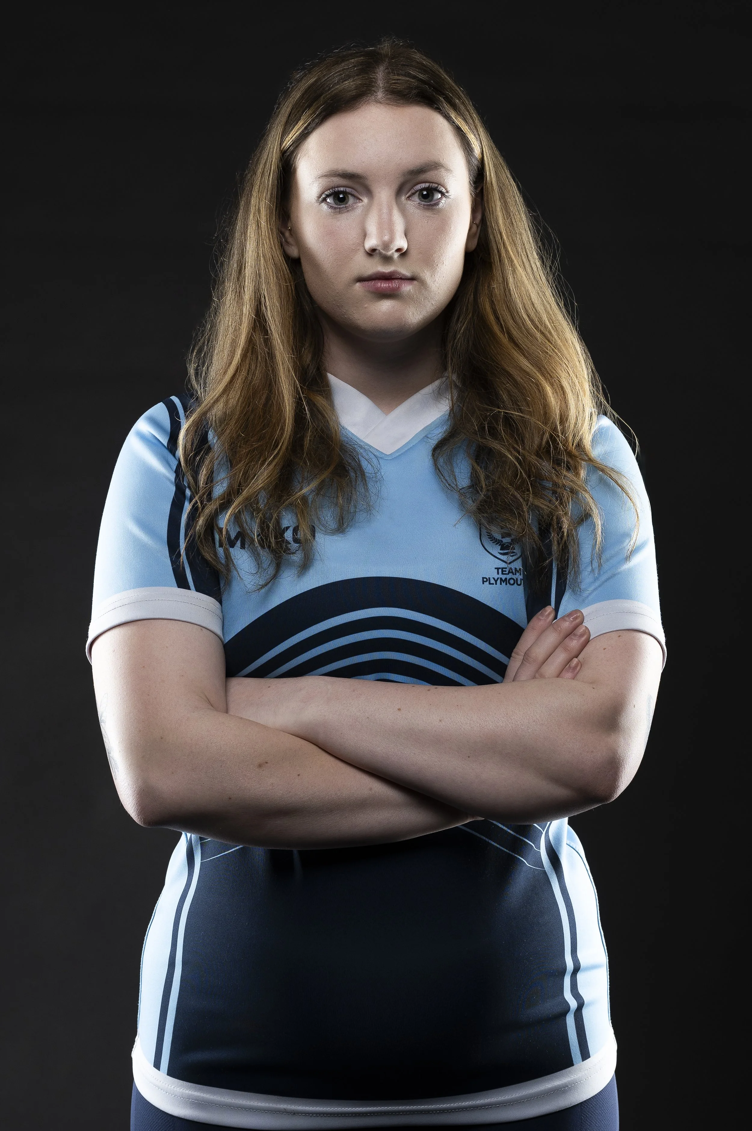 A young woman with long red hair, crossed arms, wearing a light blue athletic jersey with dark blue and white accents, standing against a black background.