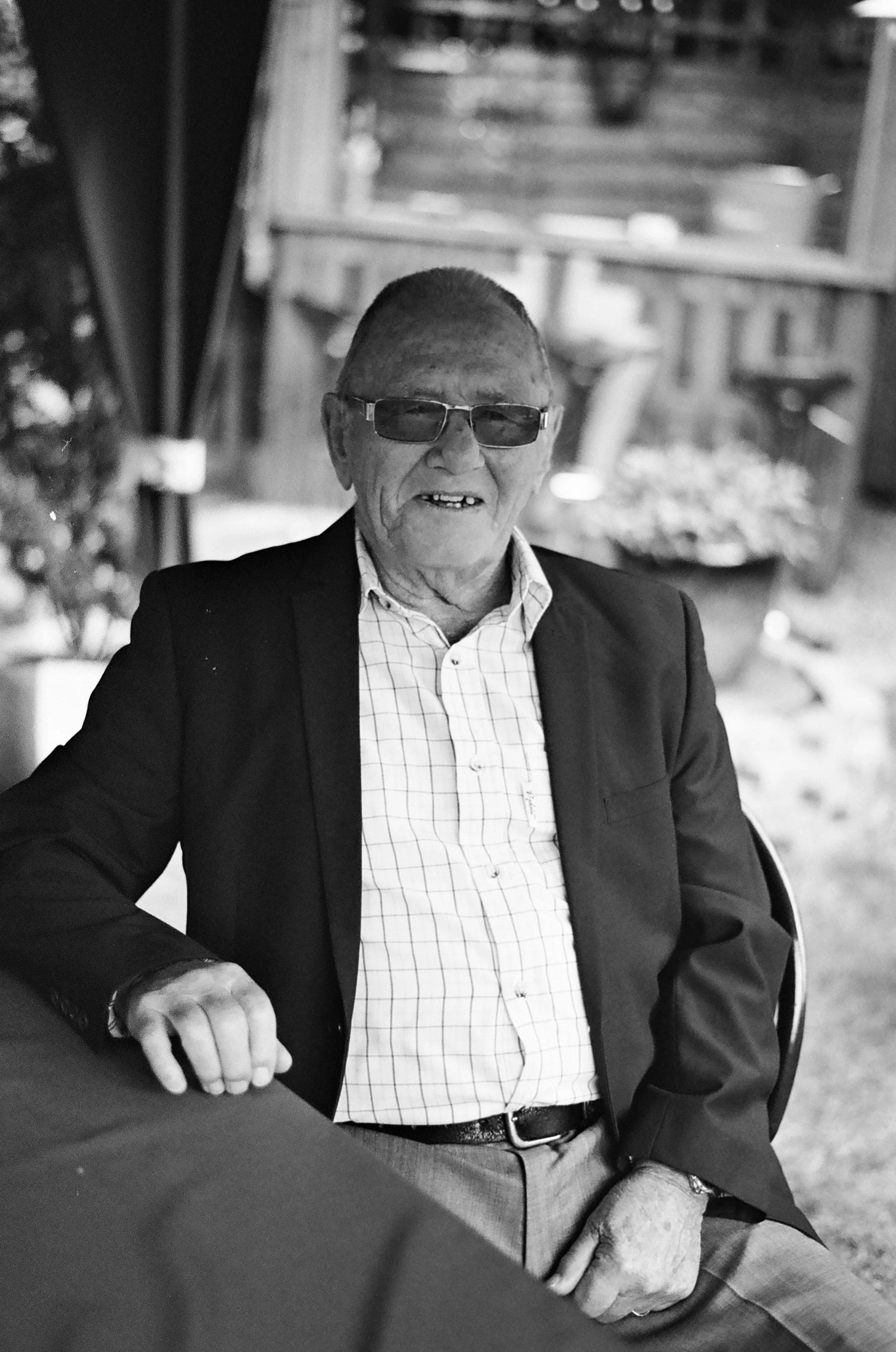 Black and white photo of an elderly man wearing a suit and glasses, sitting at a table outside.