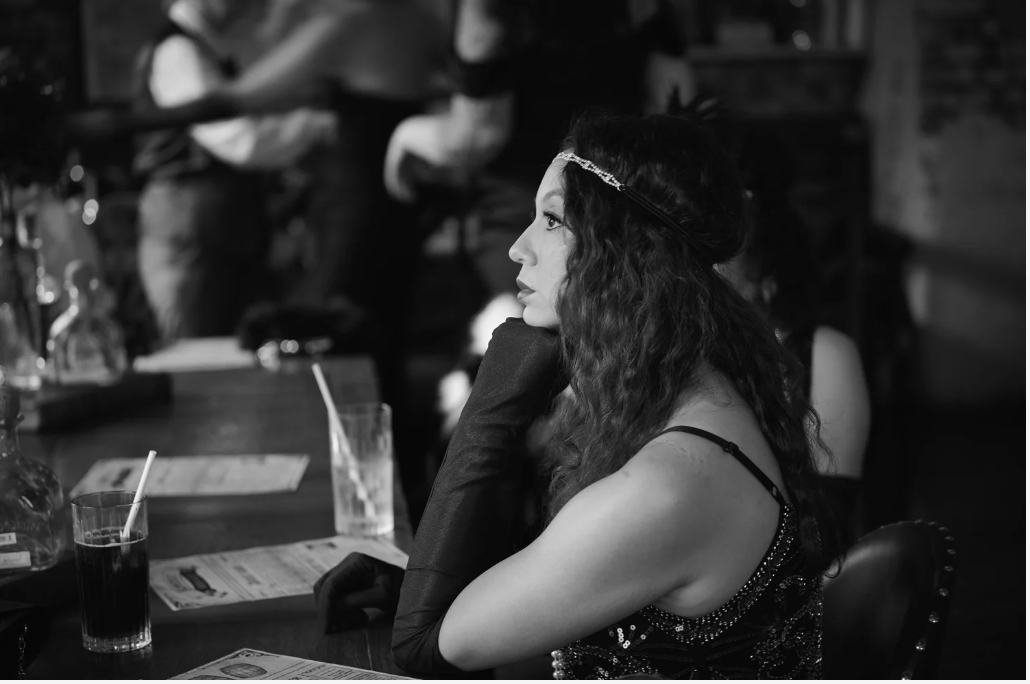 A woman with long, curly hair wearing a headband and dress, sitting at a restaurant table, resting her chin on her hand and looking to the side in black and white.