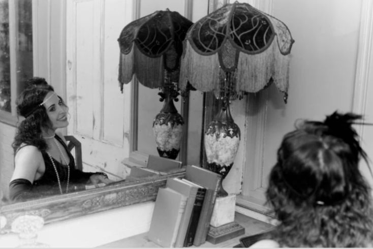 A woman dressed in period costume looking at her reflection in a mirror with two ornate table lamps on a wooden vanity.