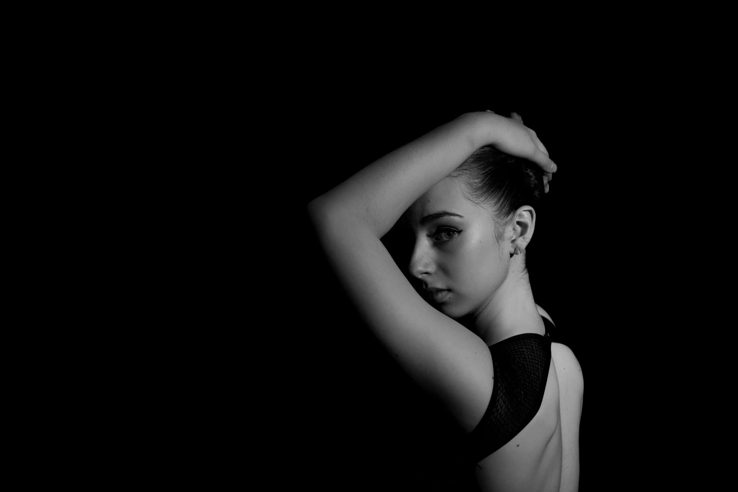 Black and white portrait of a person with arm raised, looking over shoulder against a dark background.
