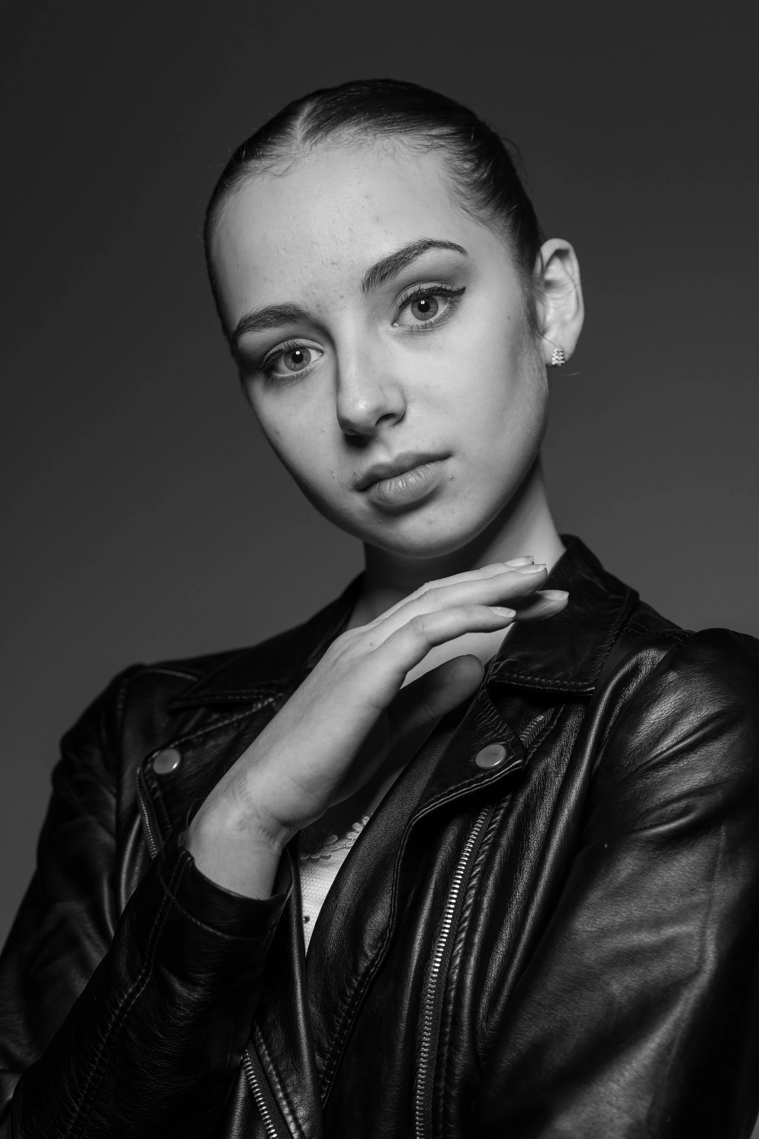 Black and white portrait of a person wearing a leather jacket, with a serious expression and hand raised delicately near the face.