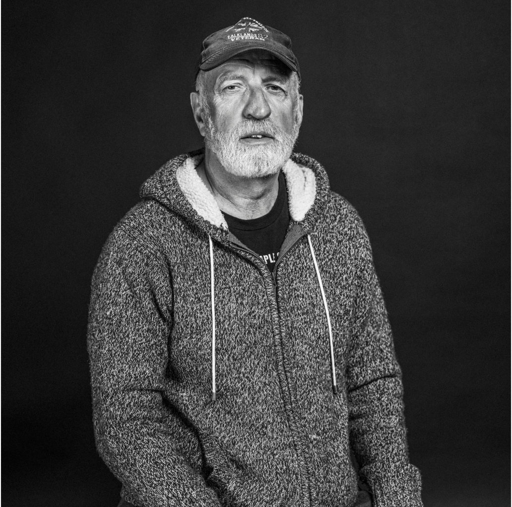 Black and white portrait of an older man with a beard and mustache, wearing a baseball cap and a zip-up hoodie with a textured pattern, standing against a plain dark background.