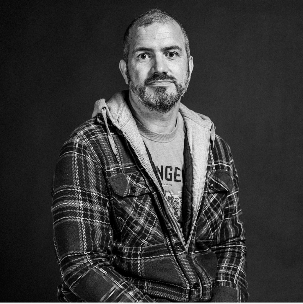 Black and white portrait of a man with a beard, wearing a plaid jacket over a hoodie and t-shirt, looking at the camera.