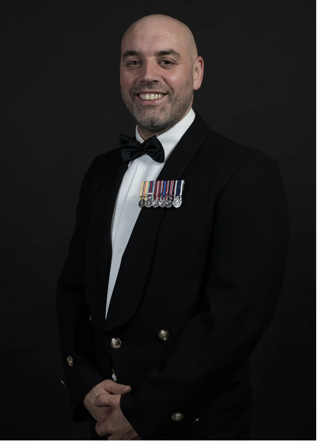 Man dressed in a formal black tuxedo with a bow tie, wearing military medals on his chest, smiling against a black background.