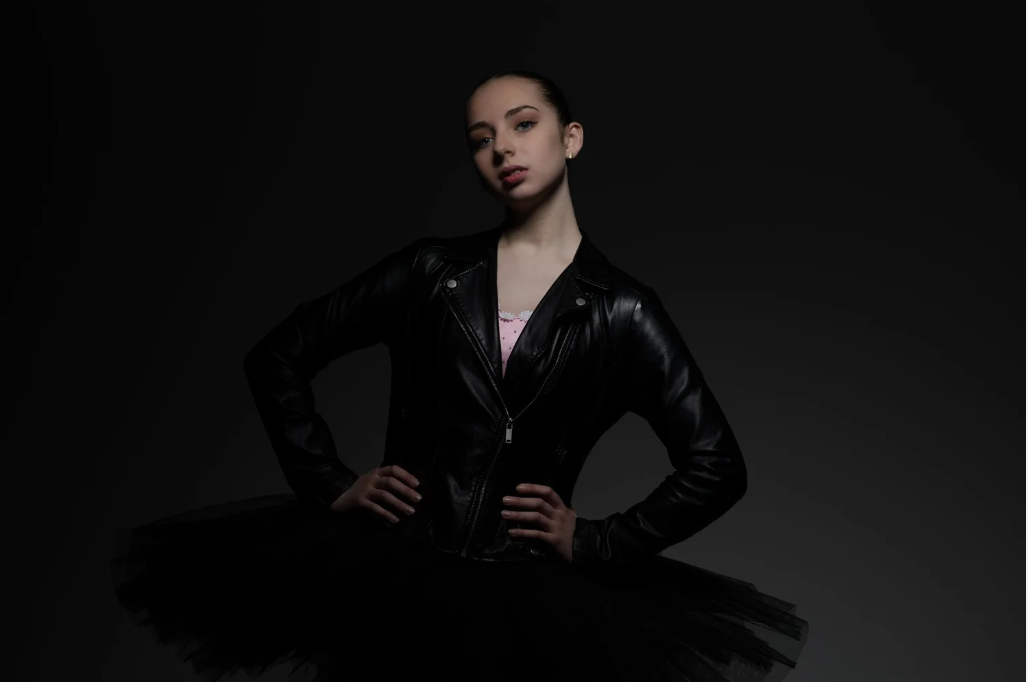 Young woman wearing a black leather jacket and black tutu skirt, posing with hands on hips against a dark background.