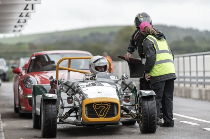 Race car driver in a vintage race car with the number 7 on the front, talking to a woman wearing a black helmet and a yellow safety vest, beside a red sports car on a racetrack.