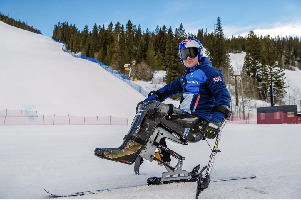 A person in a blue winter jacket and helmet using a sit-ski on snow at a ski resort, with a forested mountain background.
