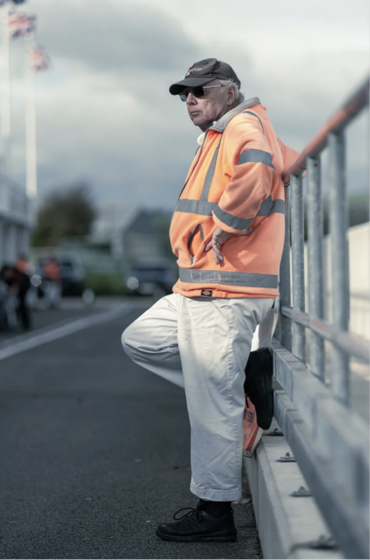 Older man in a high-visibility orange jacket and black cap leaning against a metal railing outdoors on a cloudy day.