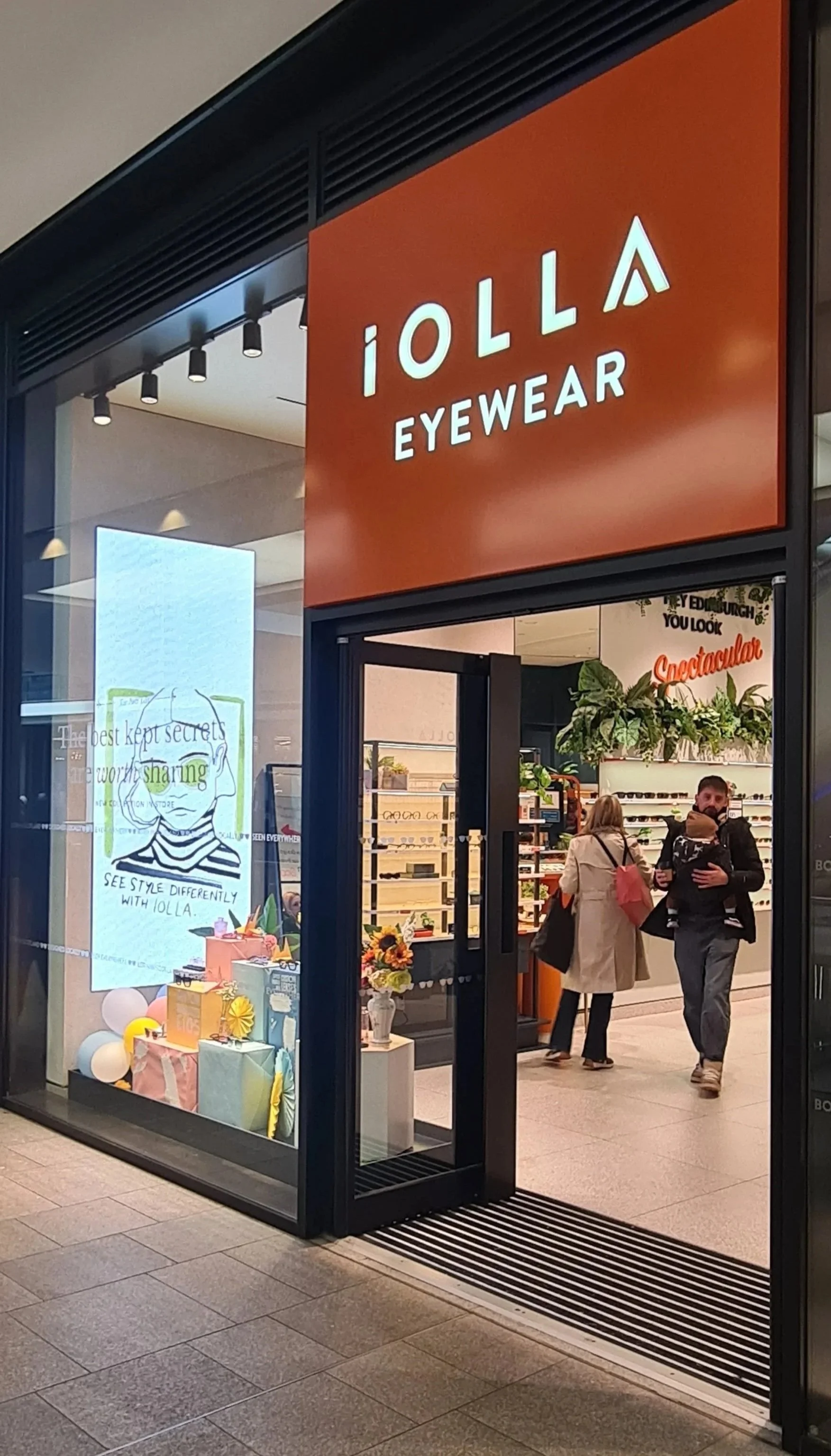 Storefront entrance of IOLLA EYEWEAR with an orange sign above the door, inside the store are customers shopping and shelves with eyeglasses.