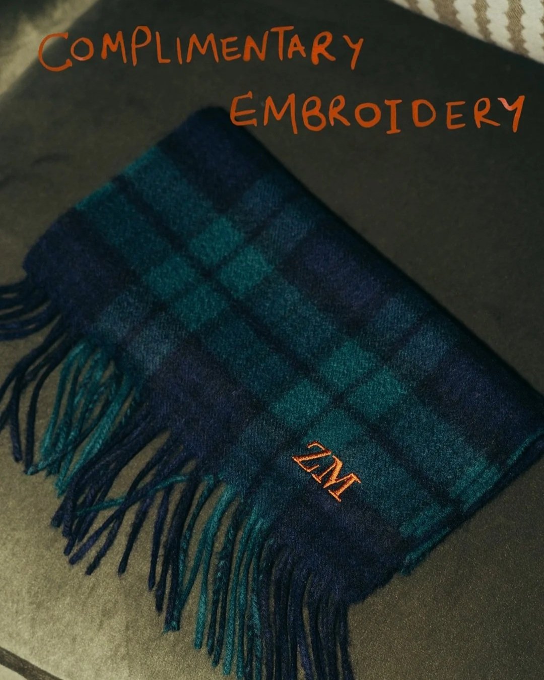 A piece of dark blue and green tartan plaid fabric with fringed edges, lying on a dark surface. The fabric has a monogram letter 'Z' embroidered in orange. Handwritten text in orange reads 'Complimentary Embroidery'.