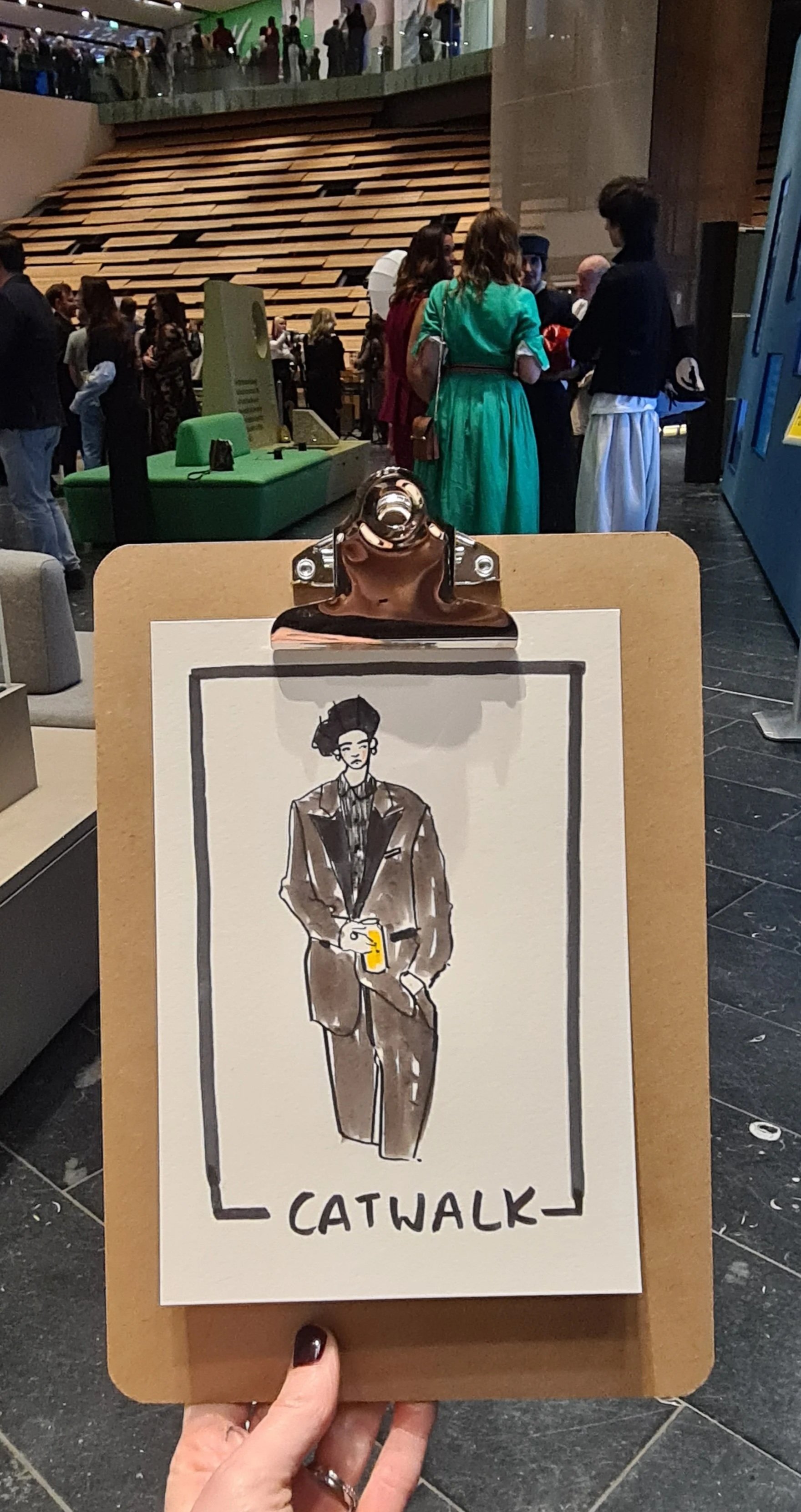A person is holding a clipboard with a drawing of a stylish man holding a drink, identified as a 'CATWALK'. In the background, people are gathered in a modern indoor space, possibly a mall or exhibition with tiered seating and a blue display on the r