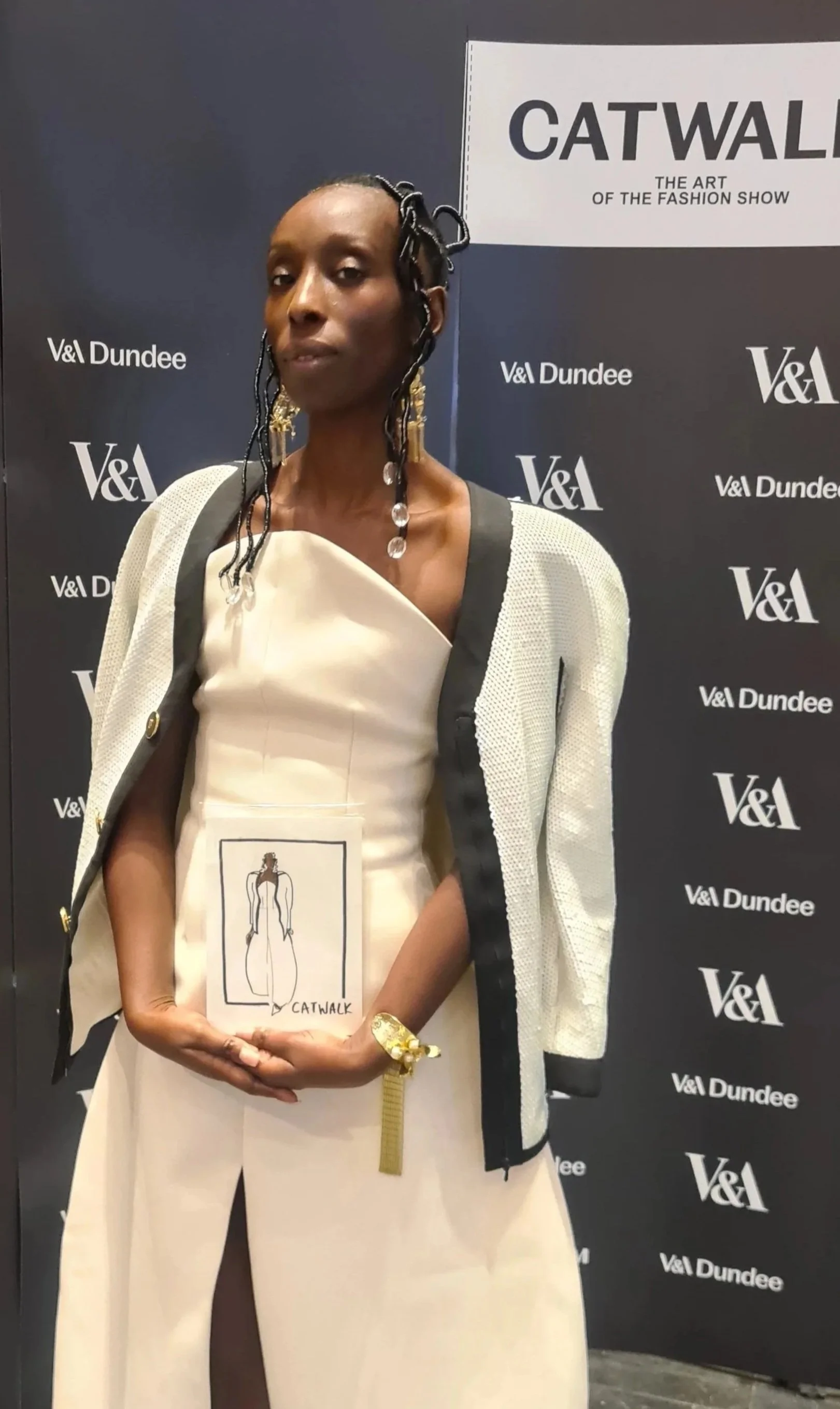 A woman at a fashion event holding a sketch of a dress. She is wearing a beige outfit with a white jacket draped over her shoulders and large earrings, standing in front of a V&A Dundee backdrop with the theme 'Catwalk: The Art of the Fashion Show'.