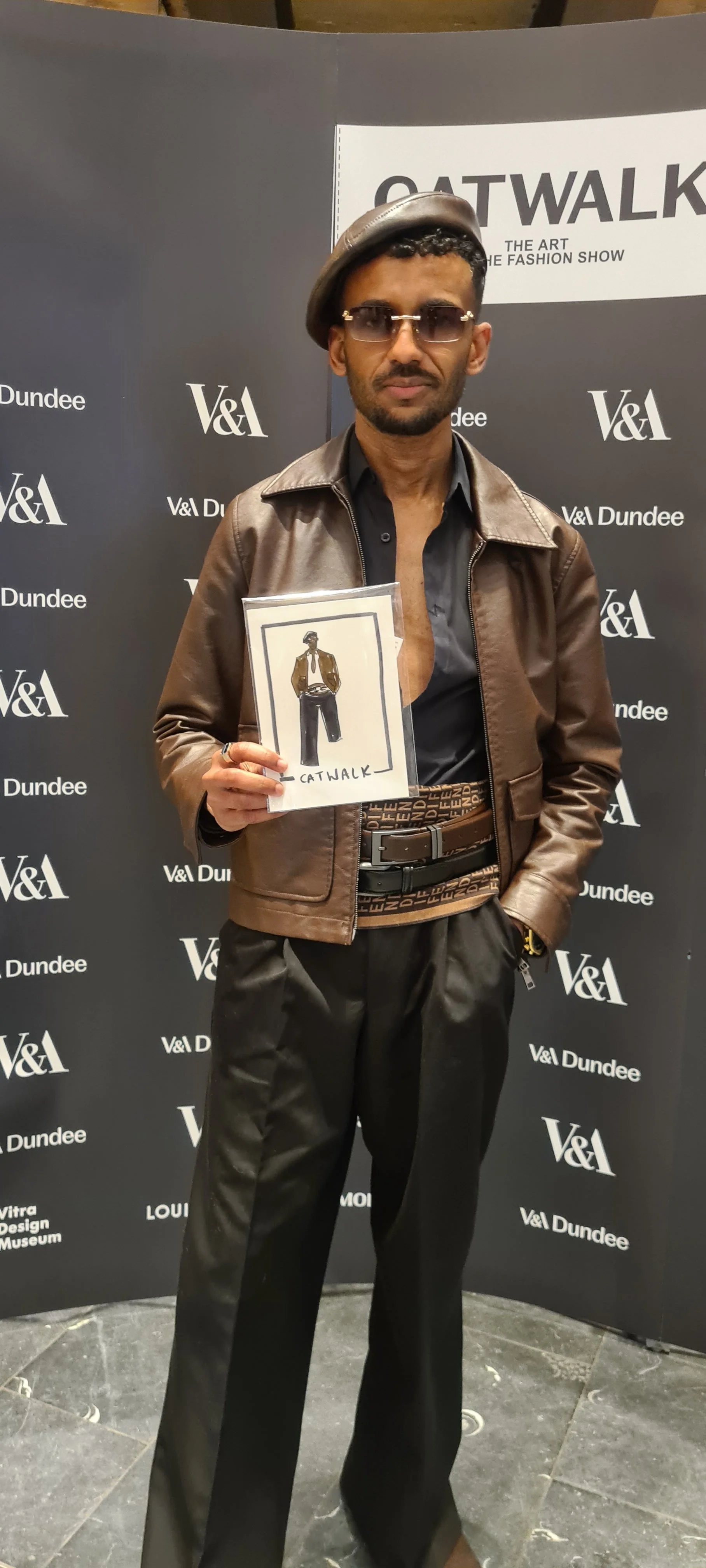 A man dressed in vintage fashion, wearing a brown leather jacket, black pants, and a gray beret, holding a sketch of a stylish outfit in front of a black backdrop with logos for V&A Dundee and an event called Catwalk.