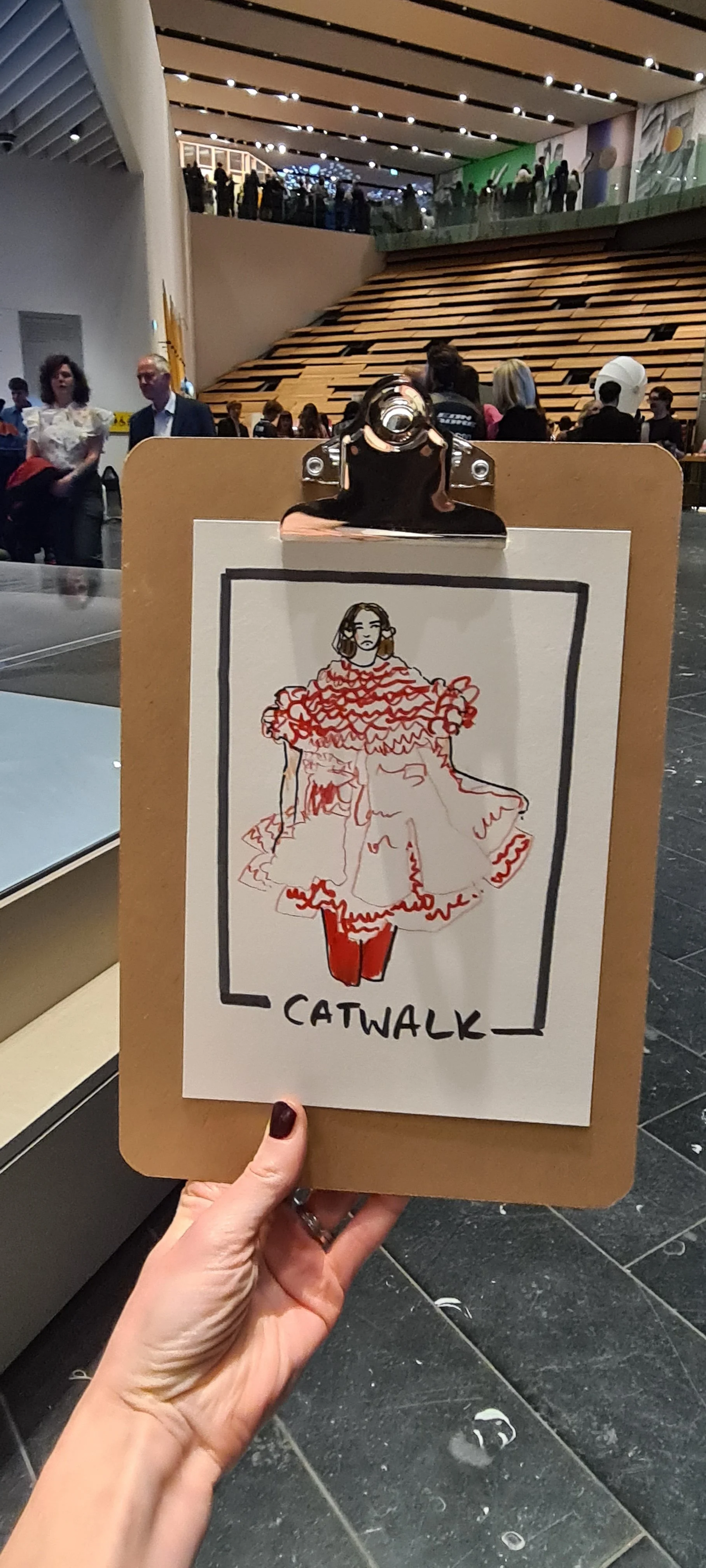 A hand holding a clipboard with a sketch of a woman wearing a detailed dress, titled 'CATWALK', in a large indoor space with people in the background.