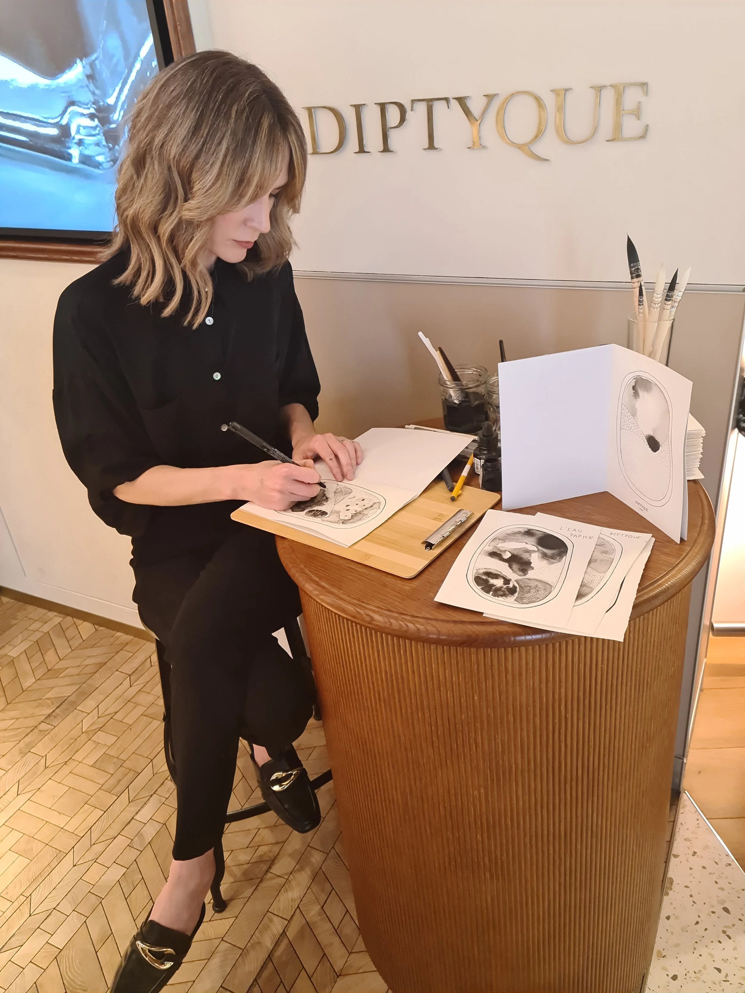 A woman with shoulder-length wavy hair, wearing a black outfit, is standing at a wooden table, drawing in a sketchbook. There are art supplies, including brushes, on the table. The background features a sign that says 'DIPTYQUE' and a modern, texture