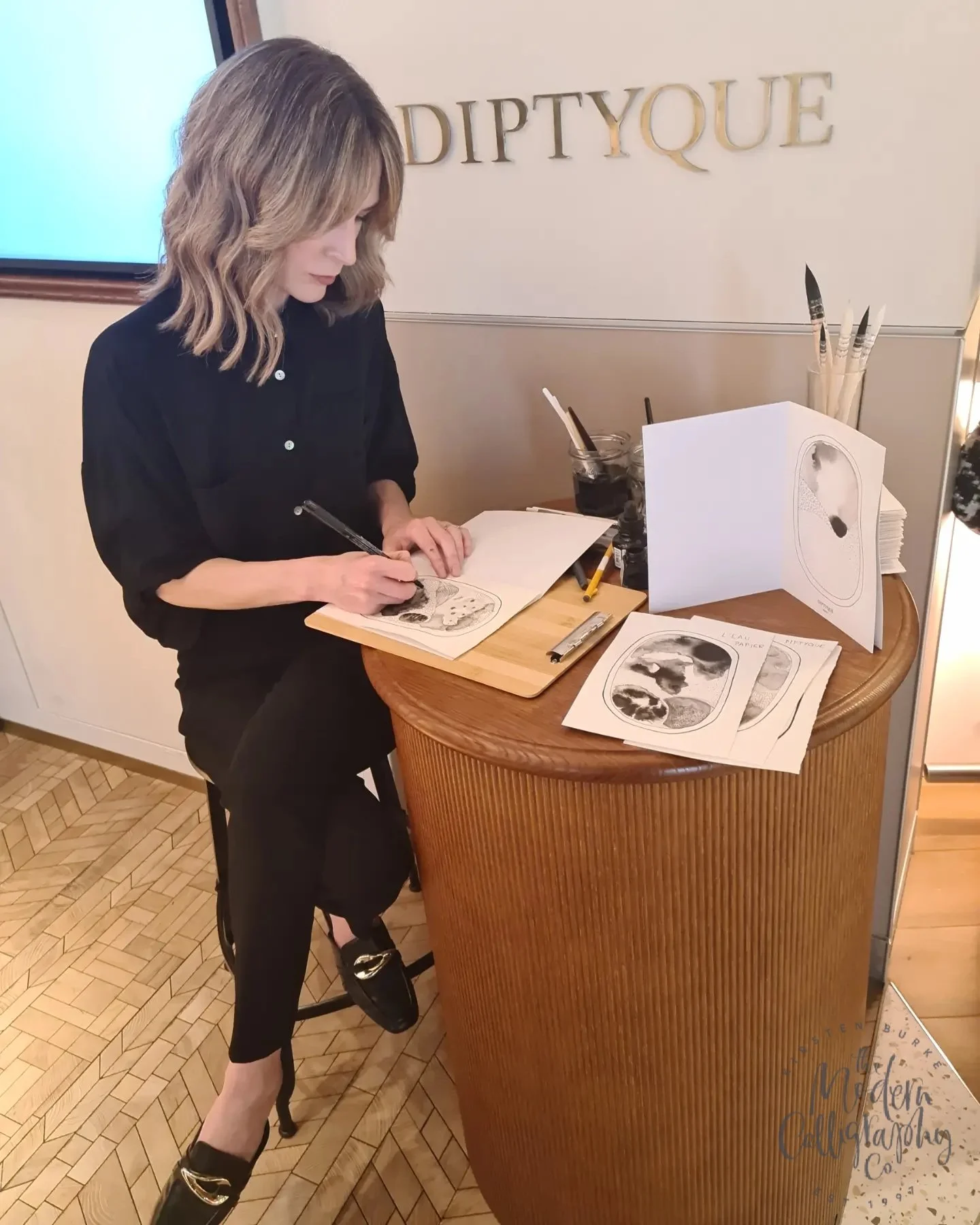 A woman sketching in a notebook at a wooden table with art supplies, including printed sketches and brushes, in a room with a 'DIPTYQUE' sign on the wall.
