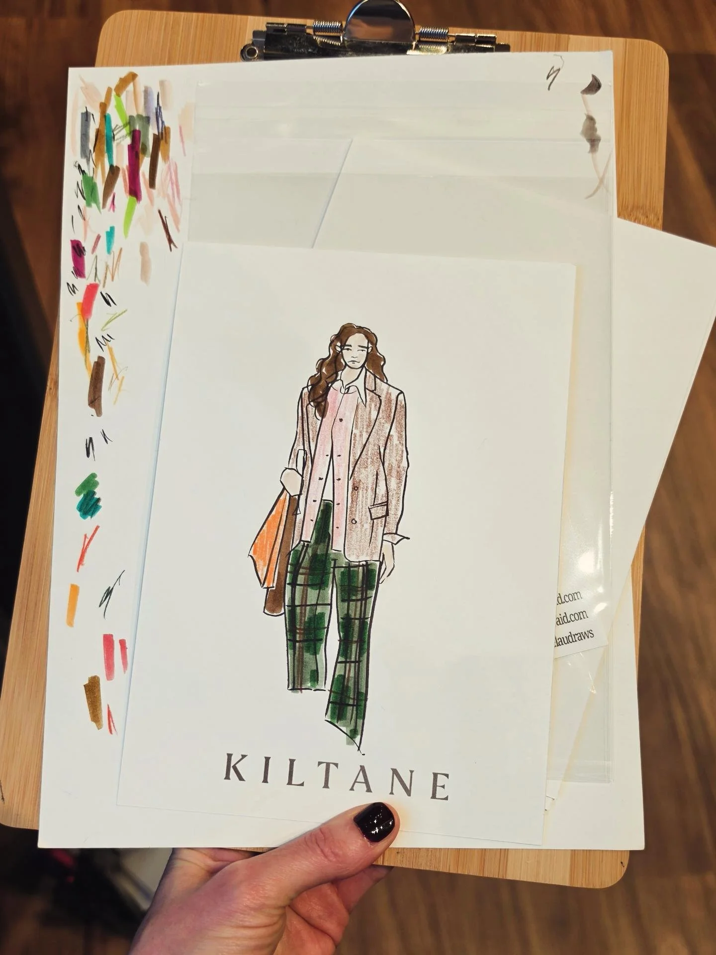A hand holding a piece of paper with a fashion illustration of a woman in a plaid suit and green plaid pants, labeled 'KILTANE'.