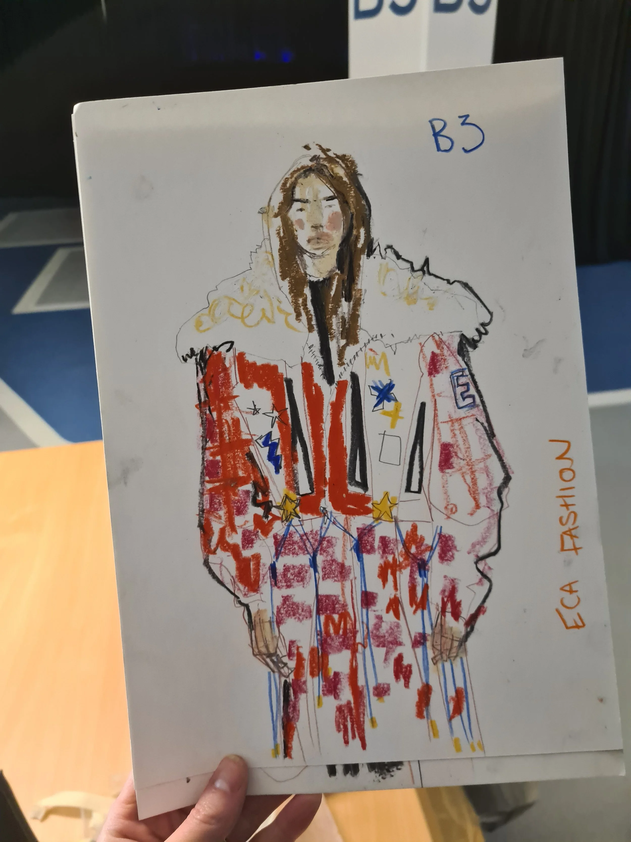 A hand holding a piece of white paper with a colorful, childlike drawing of a person wearing a red, white, and blue patriotic jacket decorated with stars and stripes. The drawing includes sketchy lines and bright colors, and the person's face is detailed with long brown hair. The paper has the words 'ELECTRIC AMERICA' and 'B3' written on it.