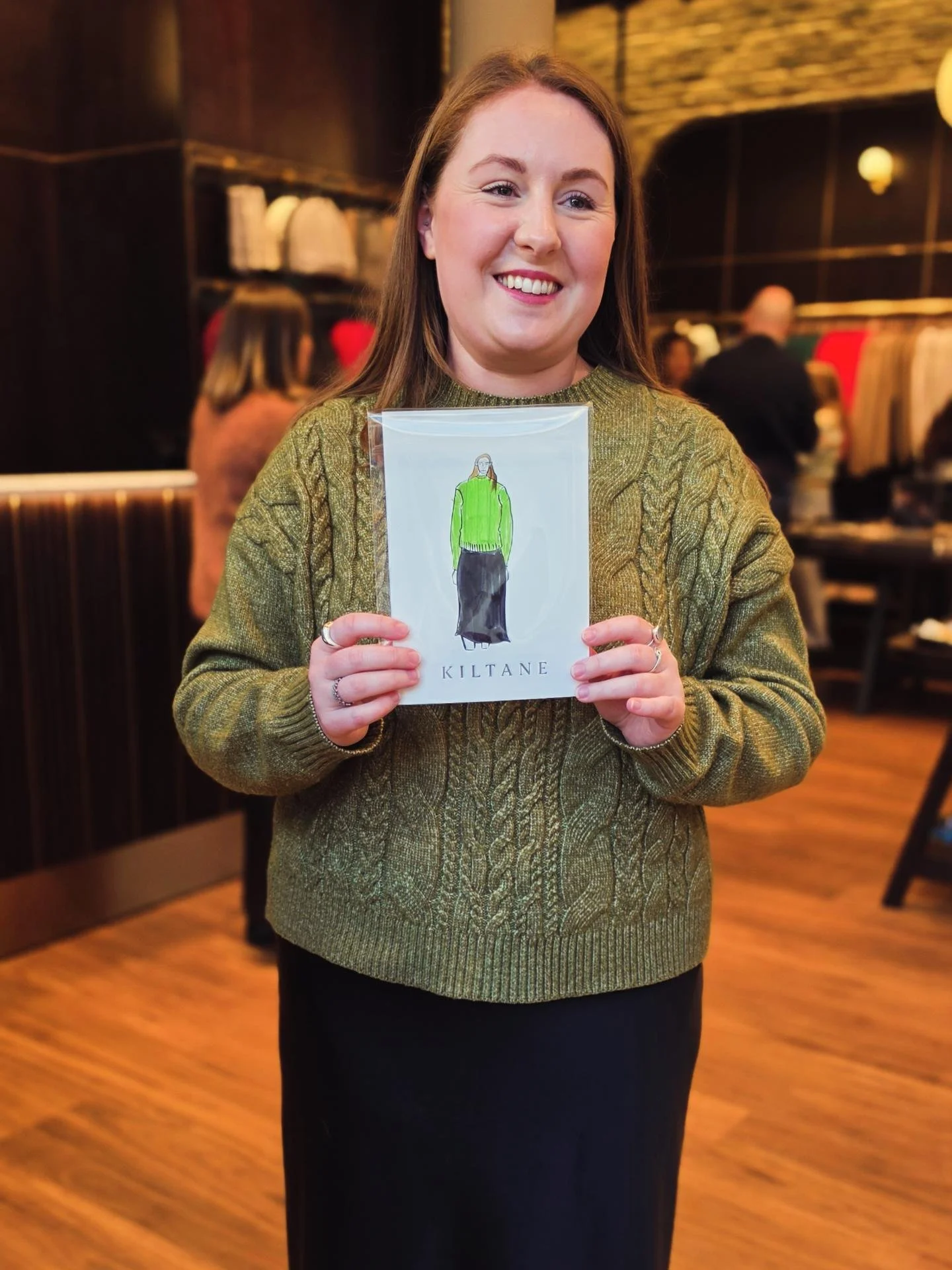 A woman with long brown hair wearing a green sweater and black pants holding a white brochure with a colorful illustration of a woman in a green sweater and black skirt, standing in a warmly lit indoor setting.
