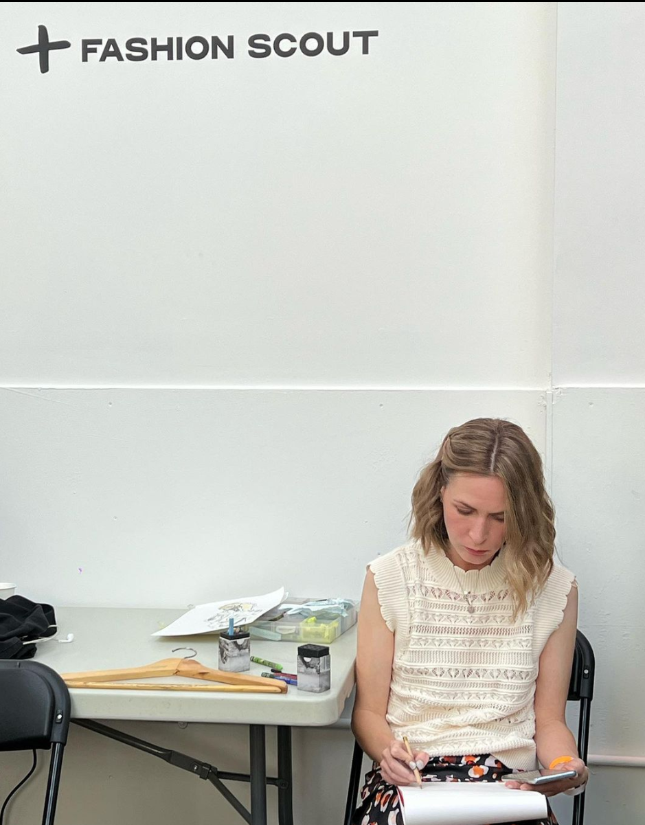 A woman sitting at a table in a room labeled 'Fashion Scout,' looking at her phone and writing on a notepad. The table has a coat hanger, some papers, and small containers.