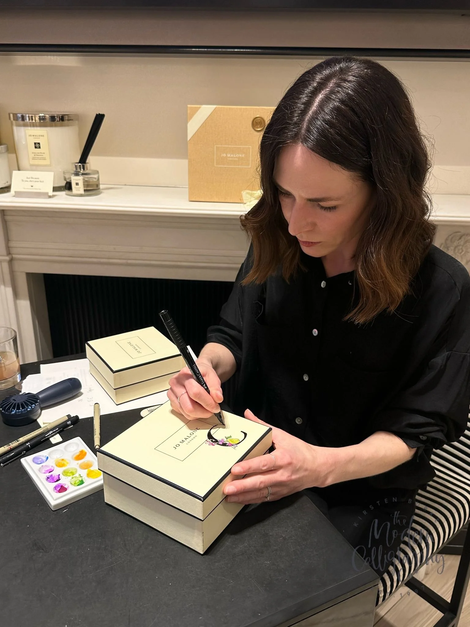 A woman with brown hair watering a cartoonish illustration of a woman and a cat wearing a pink floral dress on a piece of paper. She is sitting at a black table with a watercolor palette, black pen, and markers. There is a stack of Jo Malone gift box