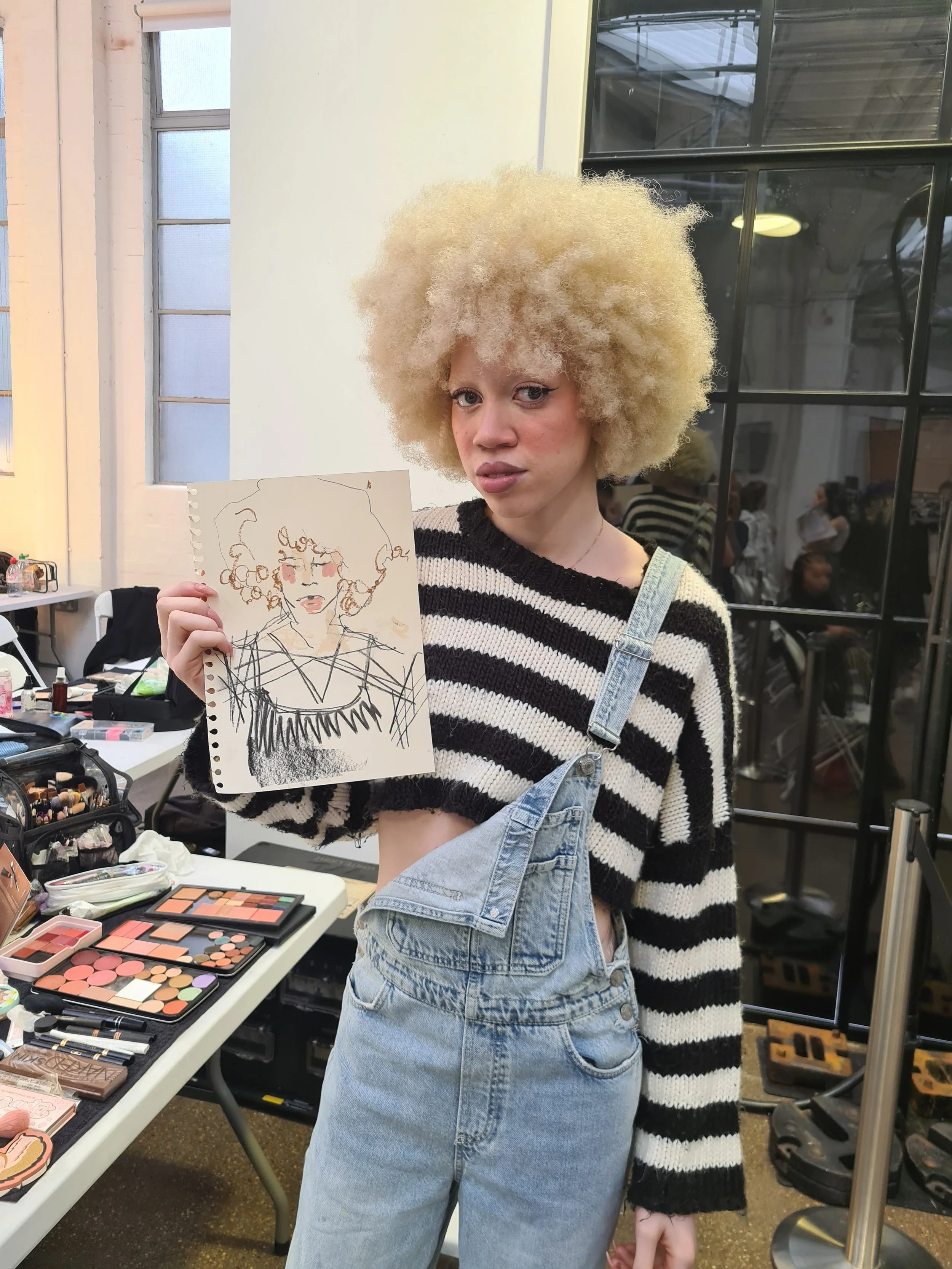 A woman with a large, curly blonde hairstyle holding a sketch-style drawing of herself, standing at a makeup table with various cosmetics, in a well-lit indoor space with large windows and a black framed glass wall in the background.