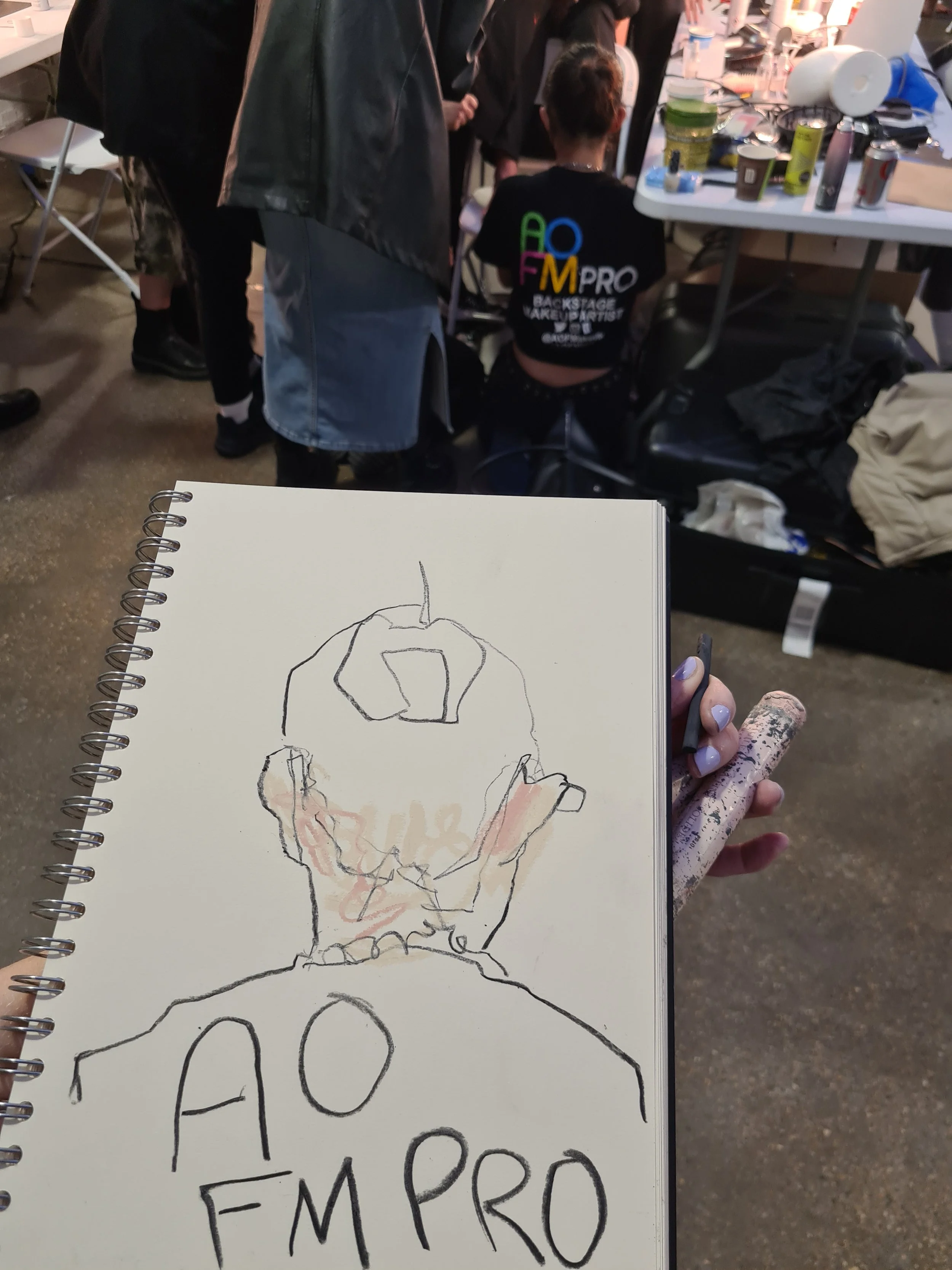 A person's sketch of a human skull on a white spiral-bound sketchpad held in their left hand, with a marker in their right hand, in front of a backstage or work area with people, tables, and various items.