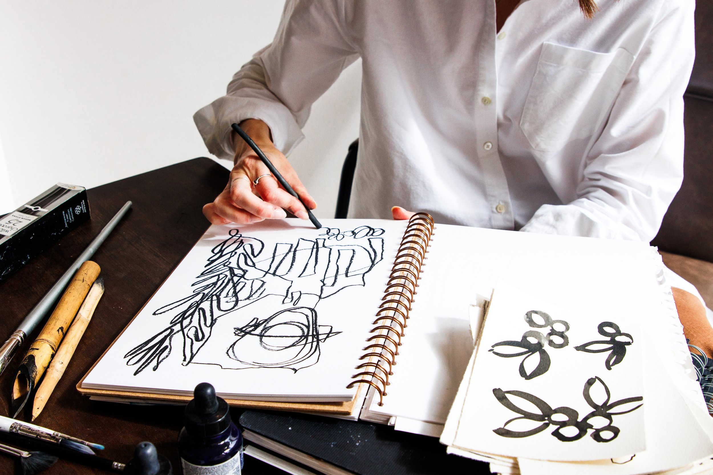 A person in a white shirt drawing abstract black ink sketches on a spiral notebook, surrounded by art supplies like brushes, markers, and ink bottles on a dark wooden table.