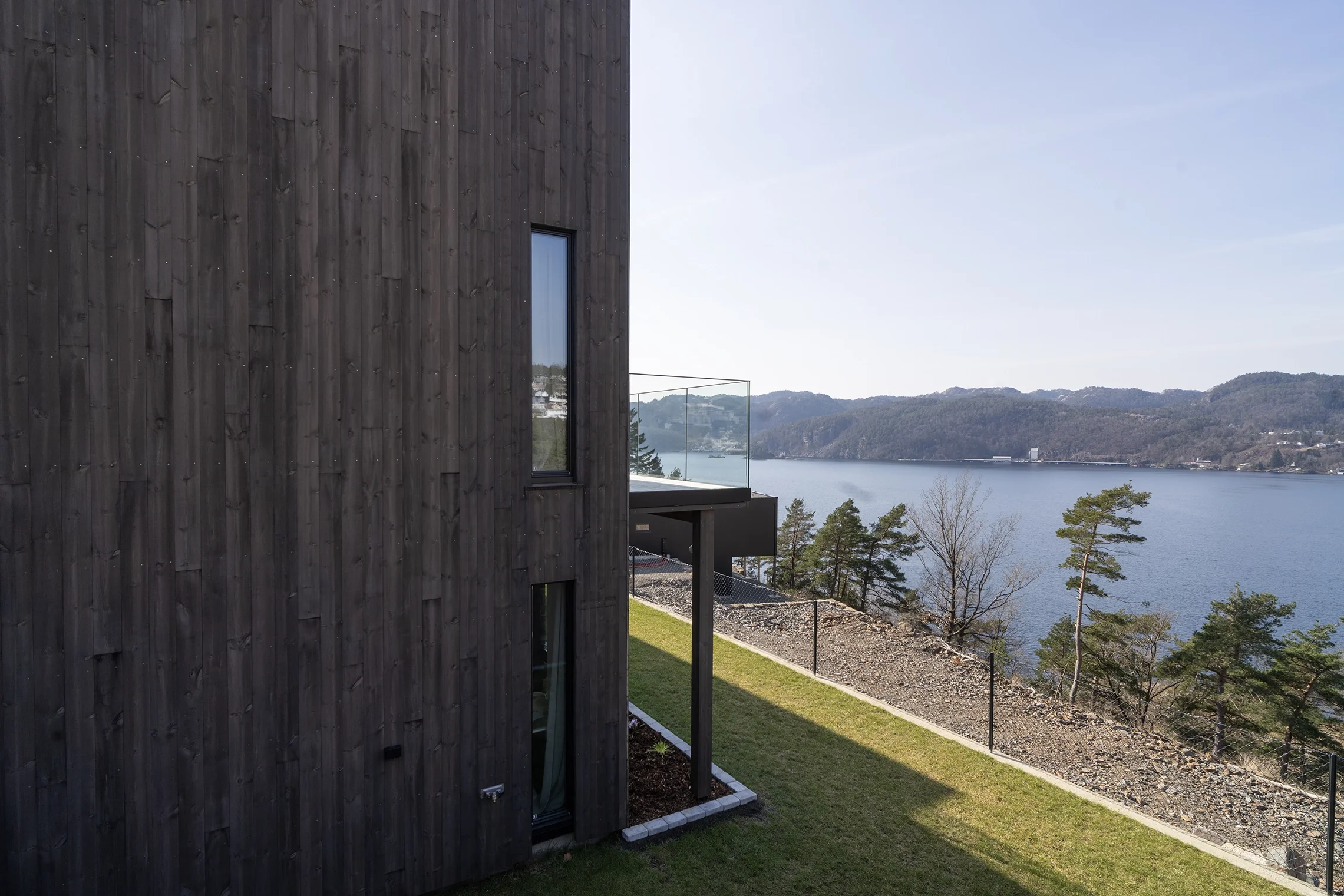 Modern house with dark wooden exterior, overlooking a lake and mountains, with a glass balcony and surrounding grassy yard.