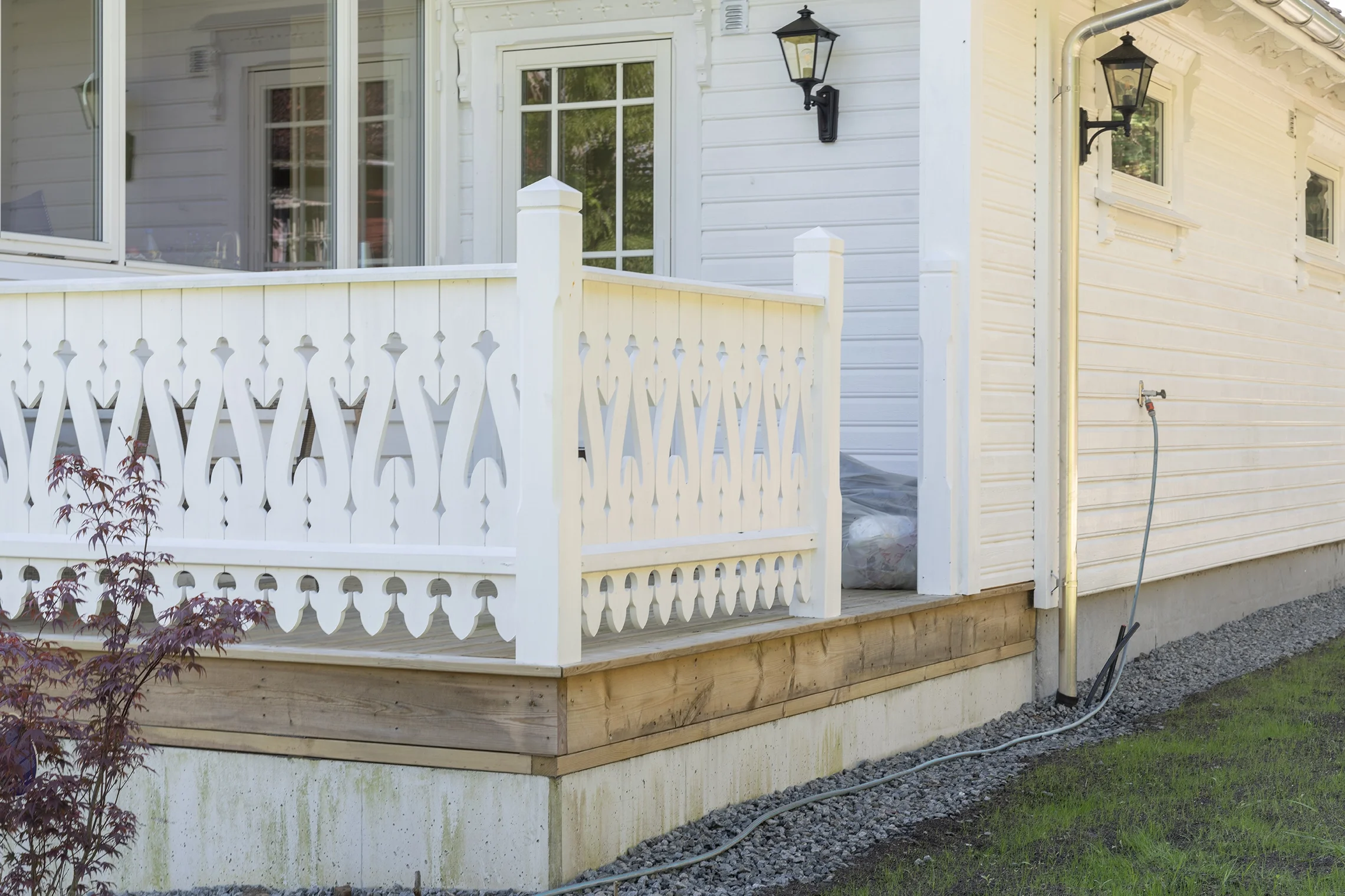 Side view of a white house with a small porch, decorative white railing, and black lantern-style exterior lights. An outdoor hose is connected to a spigot on the side of the house.