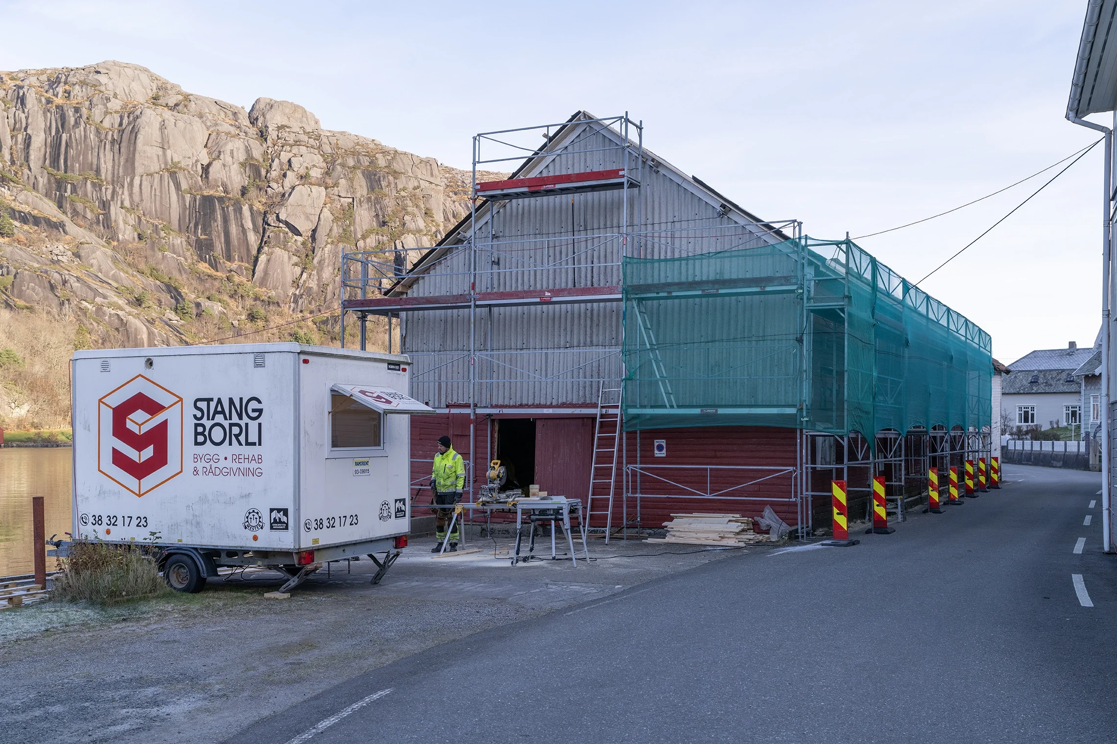 Construction site with workers and scaffolding around a barn-shaped building, a construction trailer labeled 'Stang Borli,' and a mountainous background.