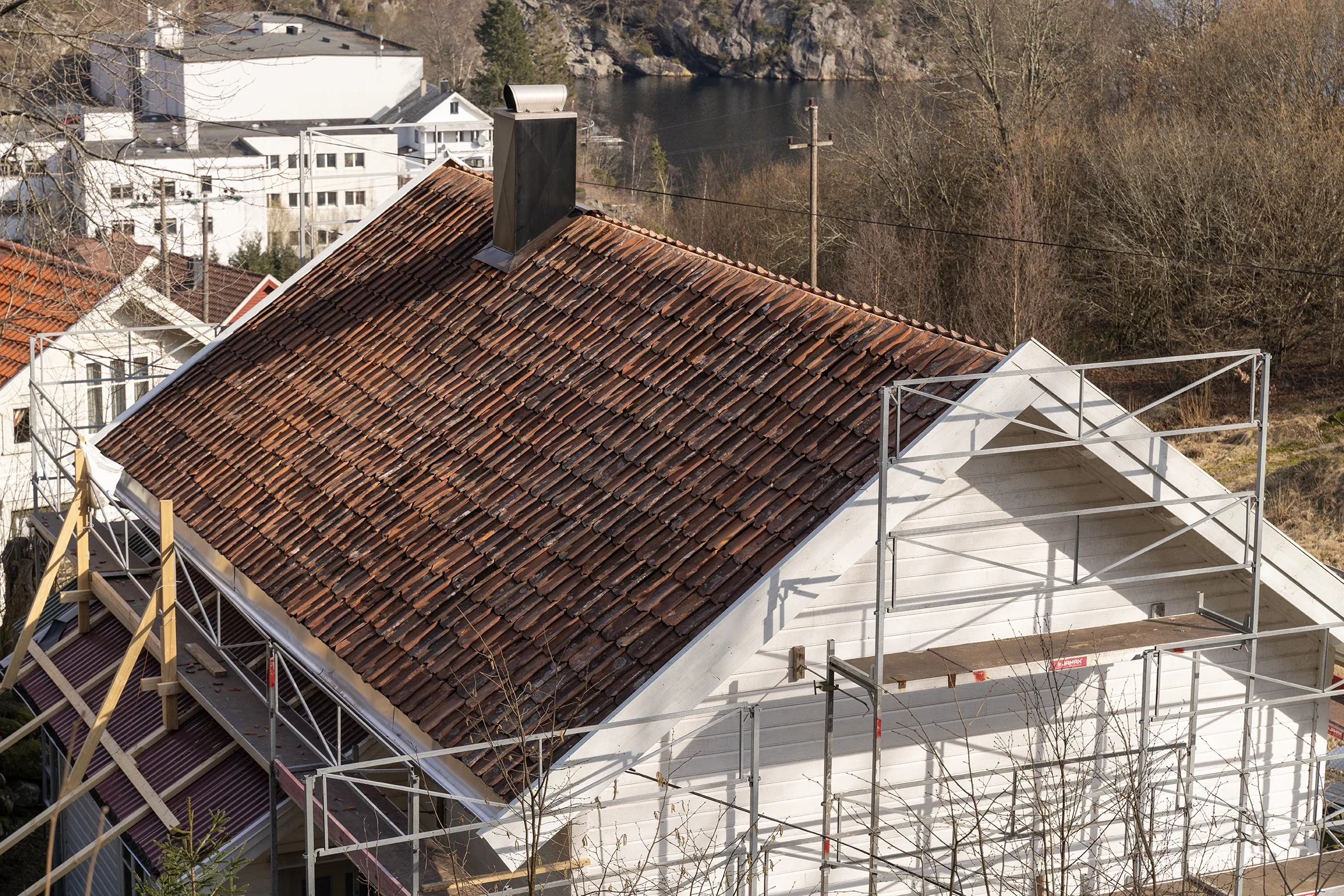 A house under construction with a sloped tiled roof, scaffolding around the structure, and neighboring buildings and trees in the background.