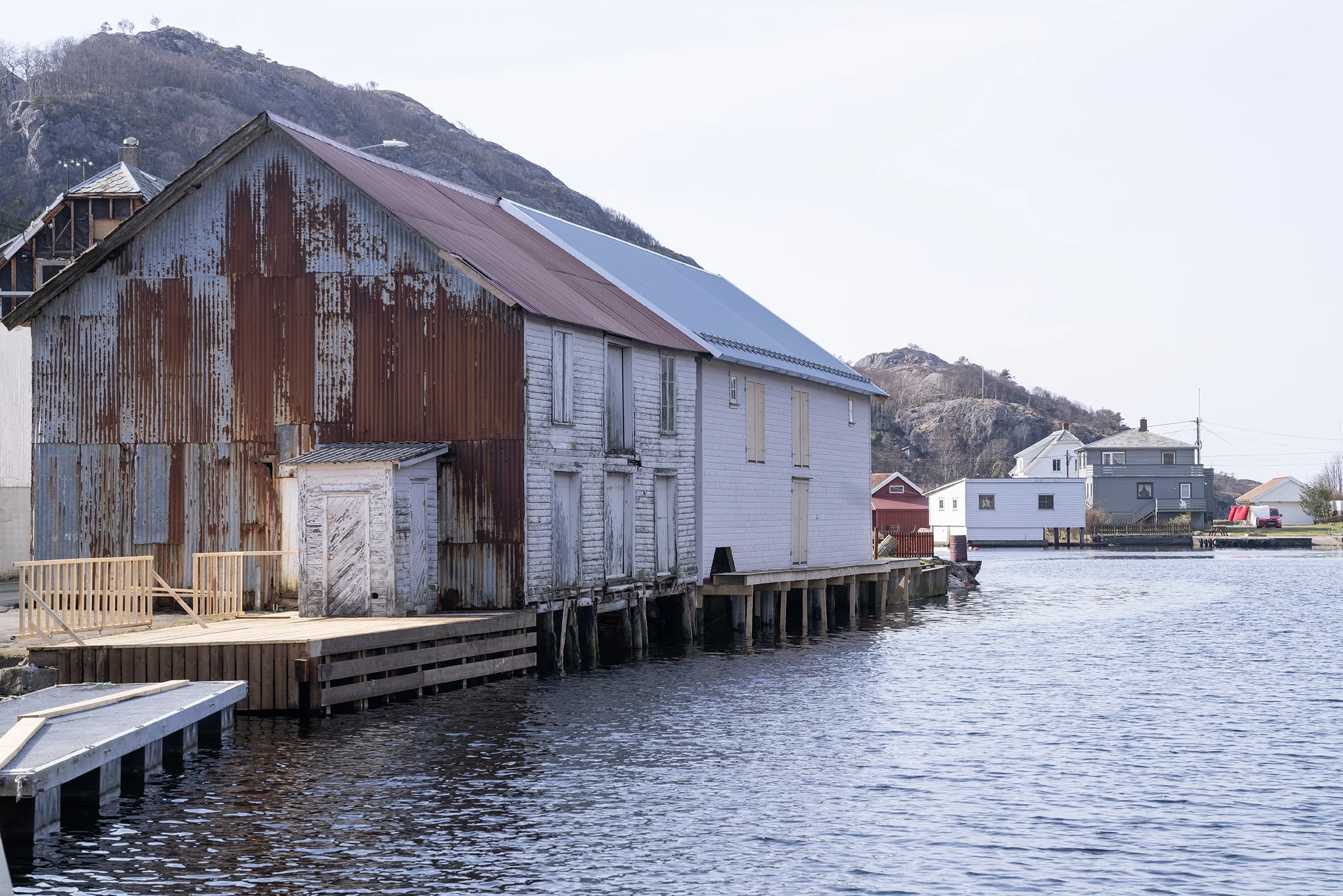 Old barn with rusted metal siding and boarded-up windows along a waterway, with houses and a mountain in the background.