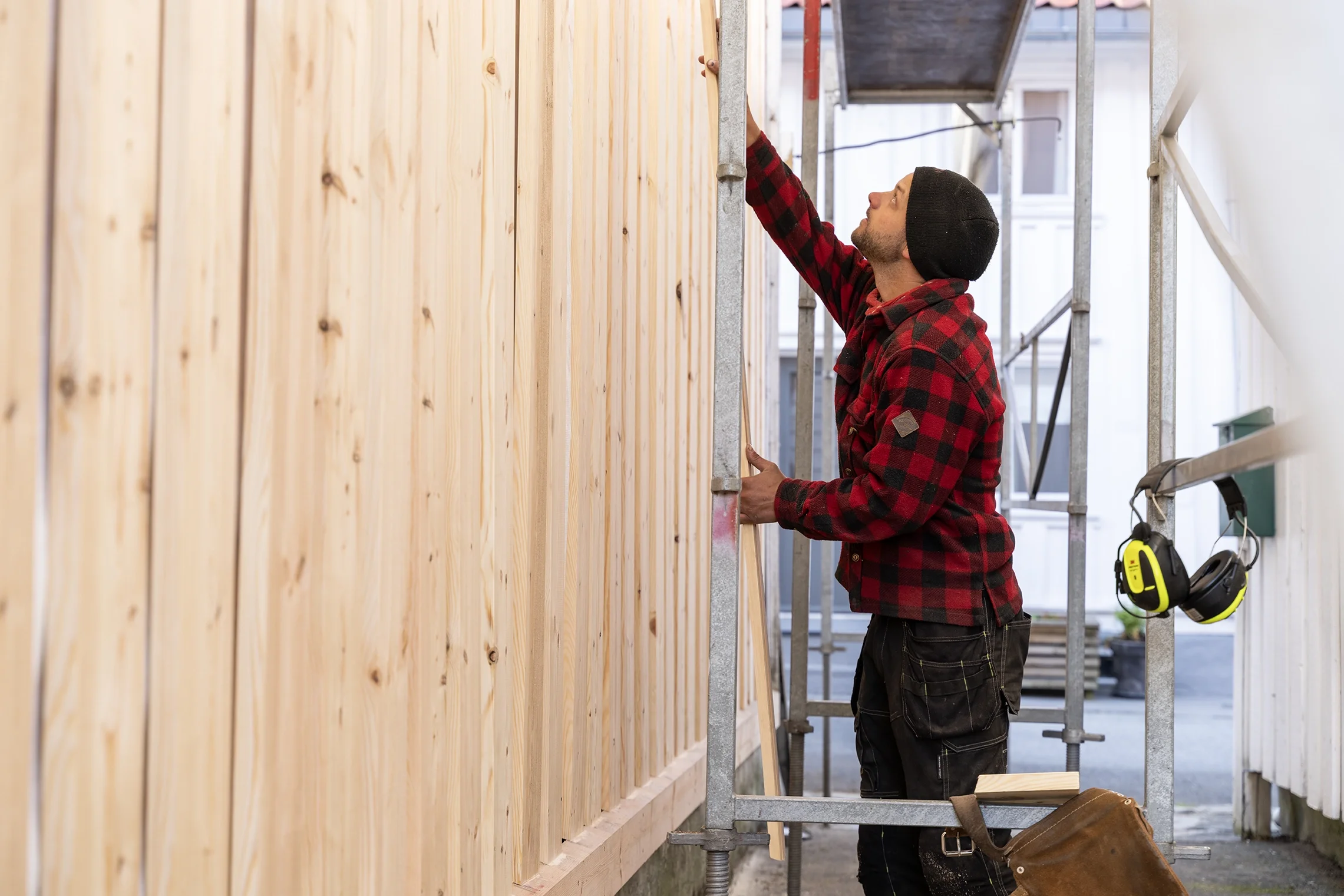 A man in a red and black checkered jacket and black beanie painting wooden planks on an outdoor scaffold.