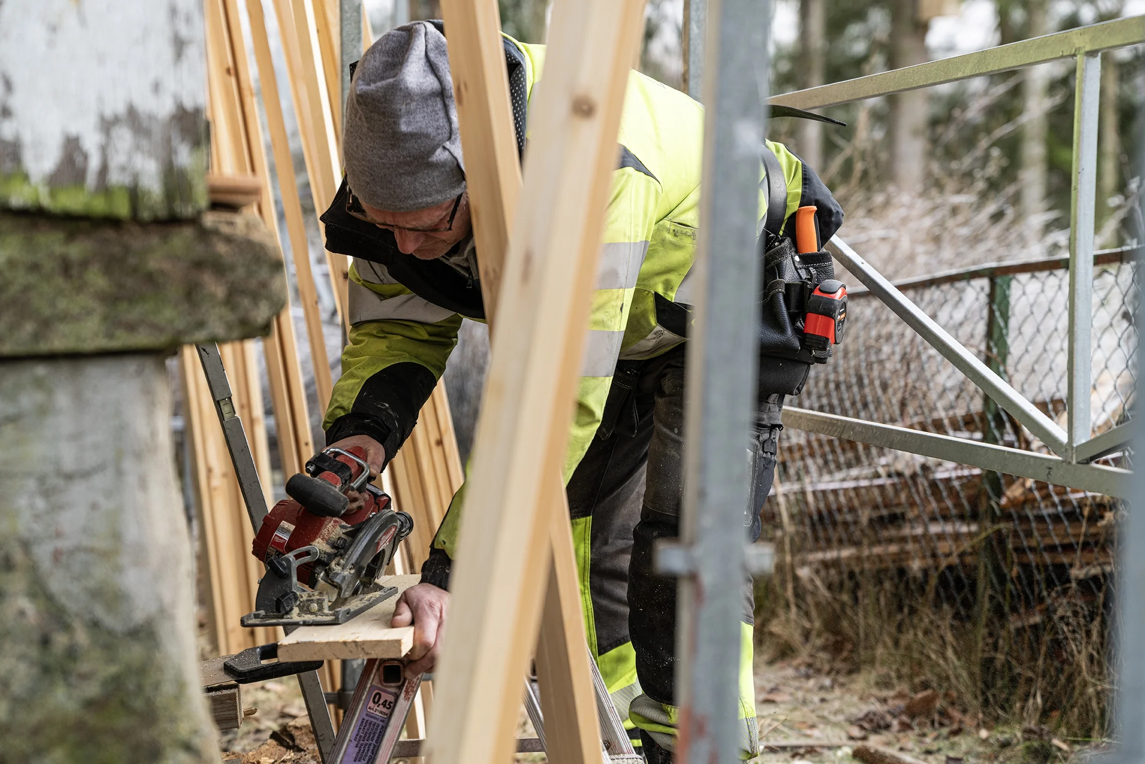 A man in a high-visibility yellow and black jacket, wearing a gray beanie and glasses, is cutting a piece of wood with a circular saw at a construction site outdoors. He is surrounded by wooden beams, metal scaffolding, and trees in the background.