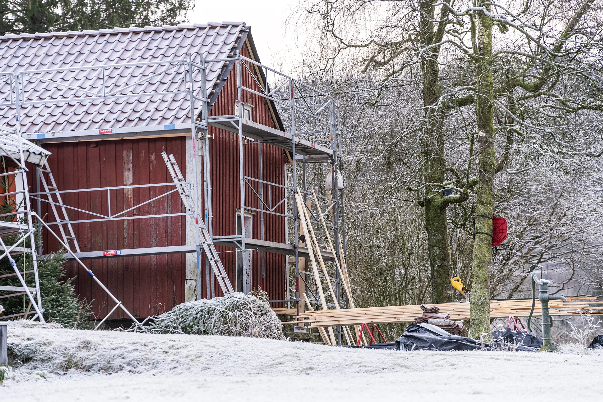 Construction of a red wooden house with scaffolding, surrounded by snow-covered trees and ground.