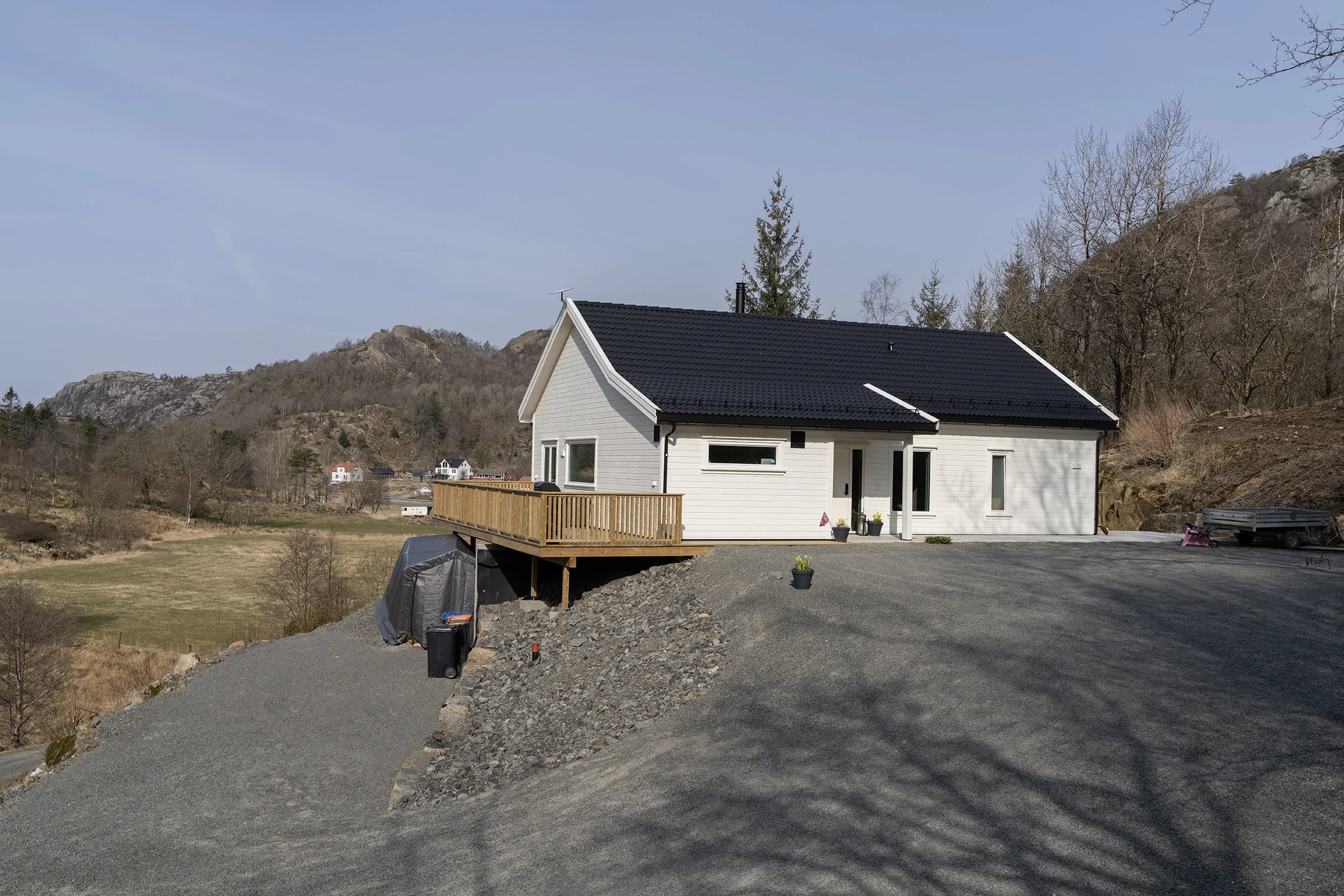 White house with black roof and wooden deck on a rocky hill, surrounded by a rural landscape with mountains and trees in the background.