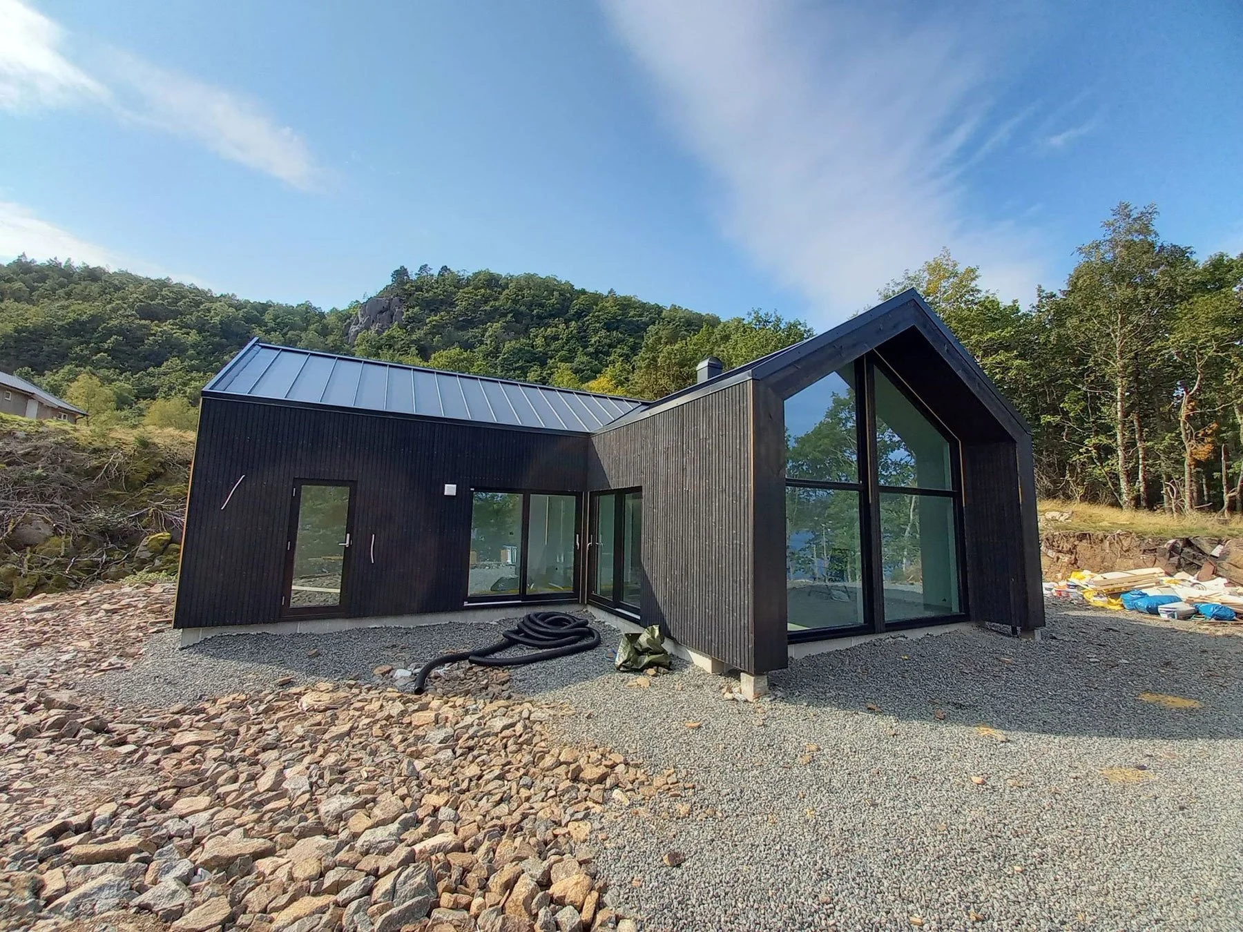 Modern black house with large glass windows and a sloped roof on a rocky terrain, surrounded by green trees and hills under a partly cloudy sky.