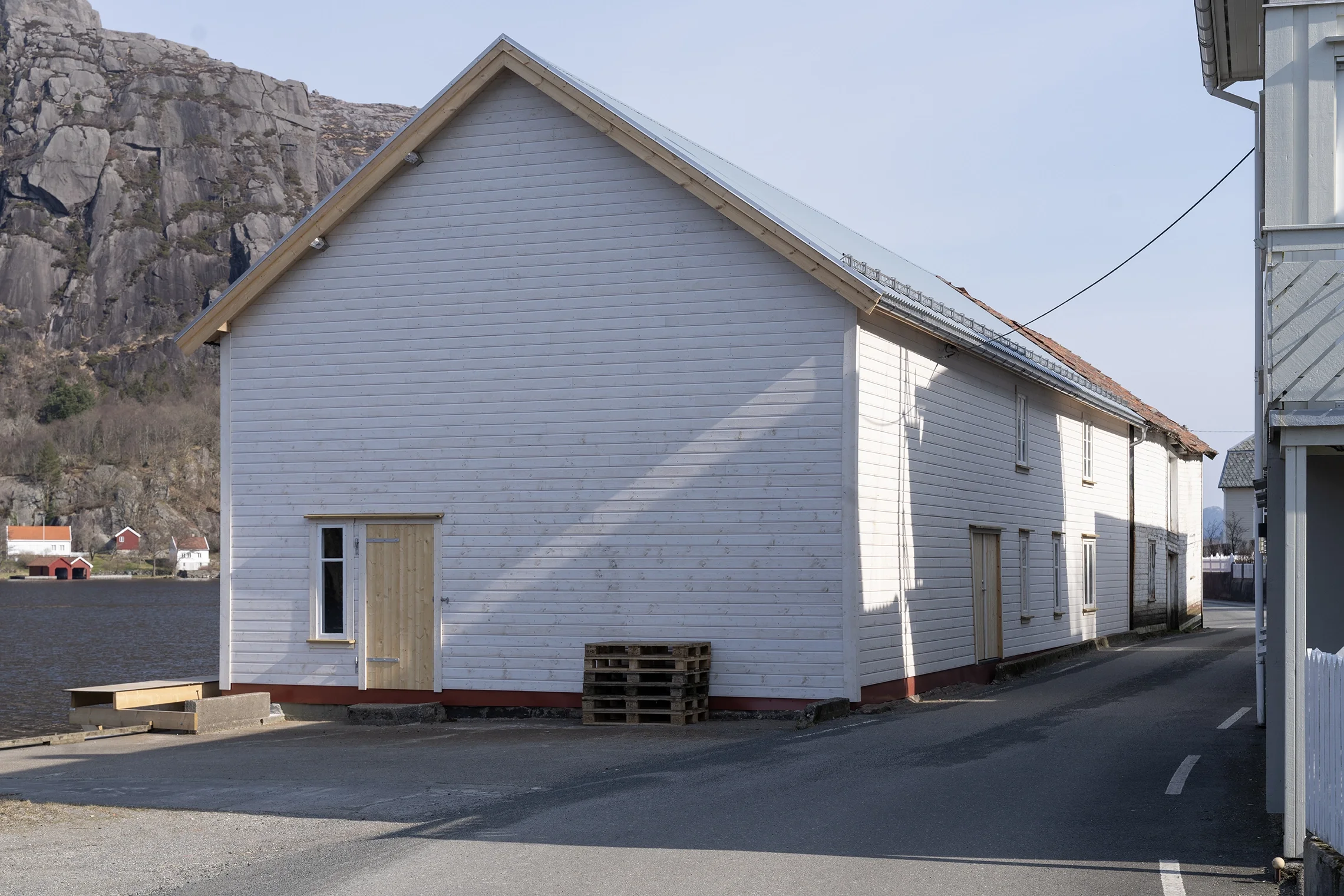 White wooden building with some windows and doors, situated near a body of water and rocky hills in the background.