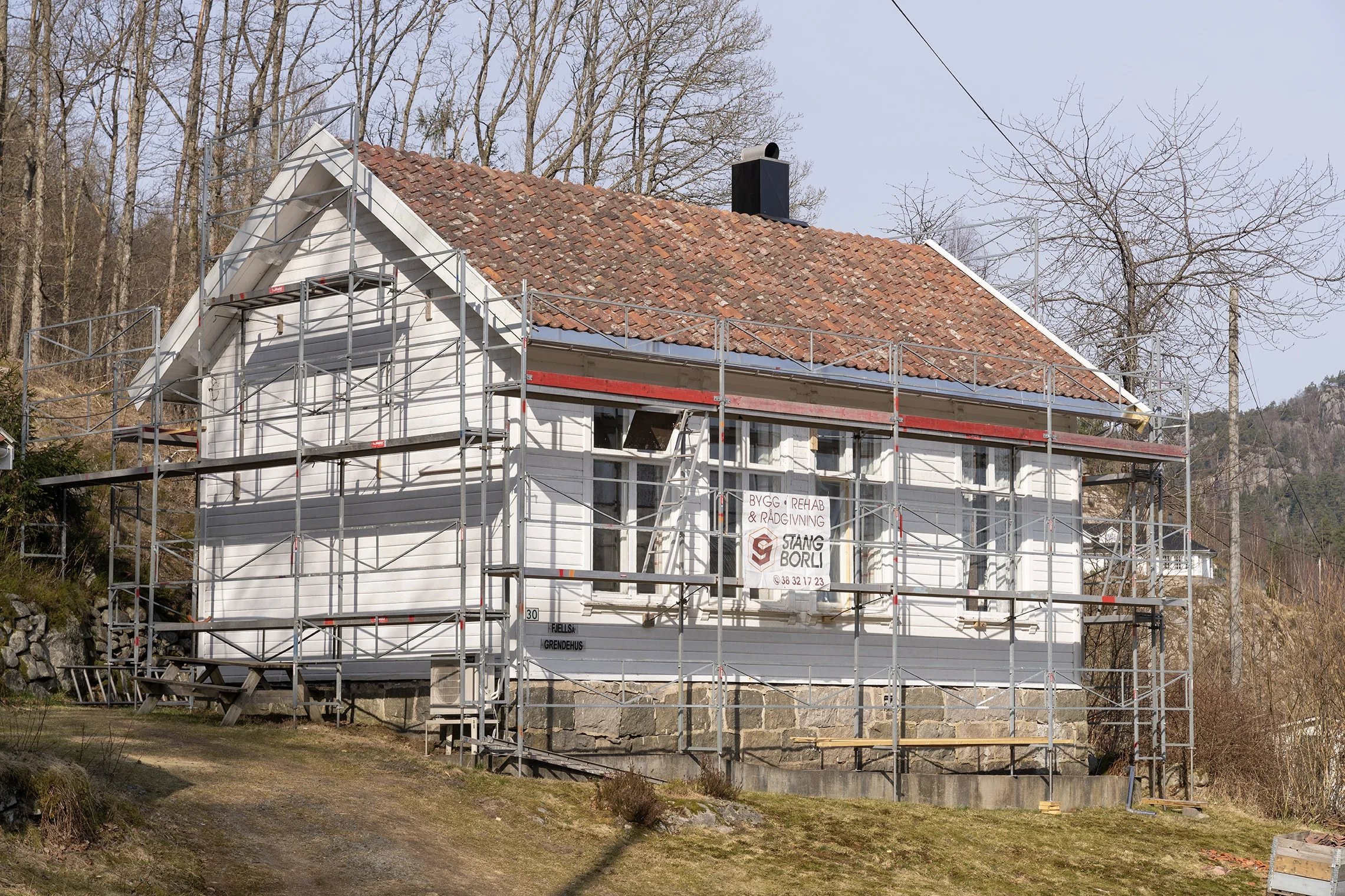 House under renovation with scaffolding surrounding it, the house has white wooden siding, a red-tiled roof, and a stone foundation.