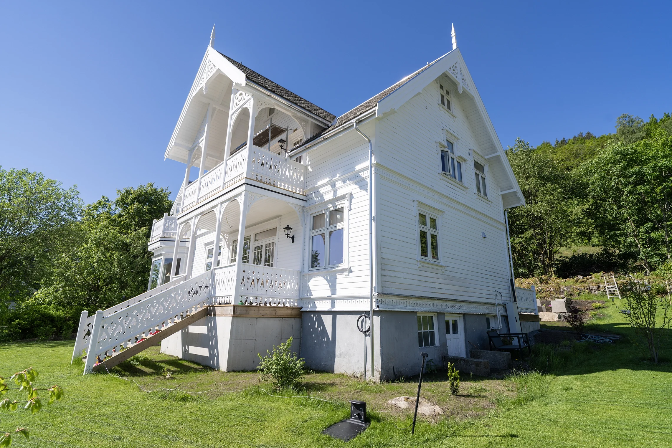 A tall, white, Victorian-style house with decorative trim, multiple stories, and a wraparound porch with intricate woodwork, set against a bright blue sky and green trees.