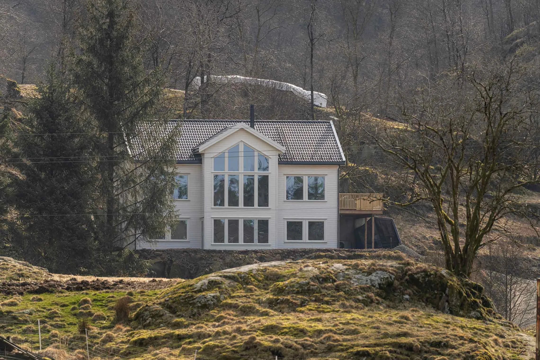 A white, multi-story house with large windows, situated on a hillside surrounded by trees and moss-covered rocks during the daytime.
