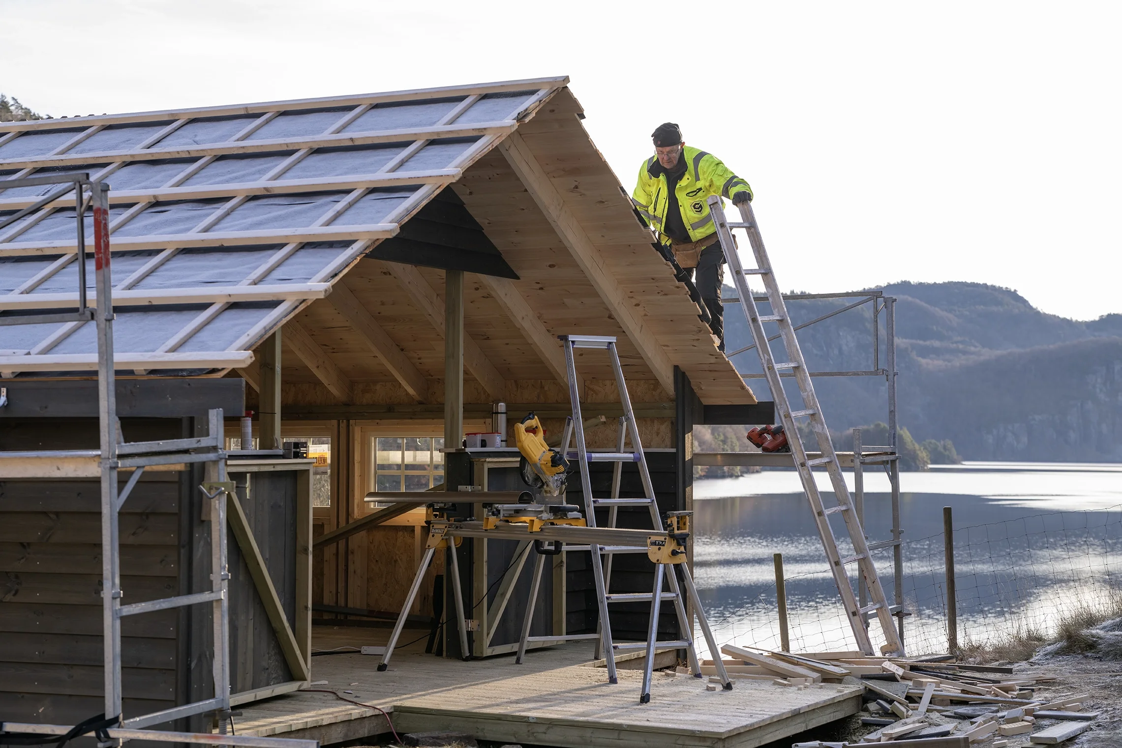 A construction worker on a ladder installing wooden roof panels on a house by a lake with mountains in the background.