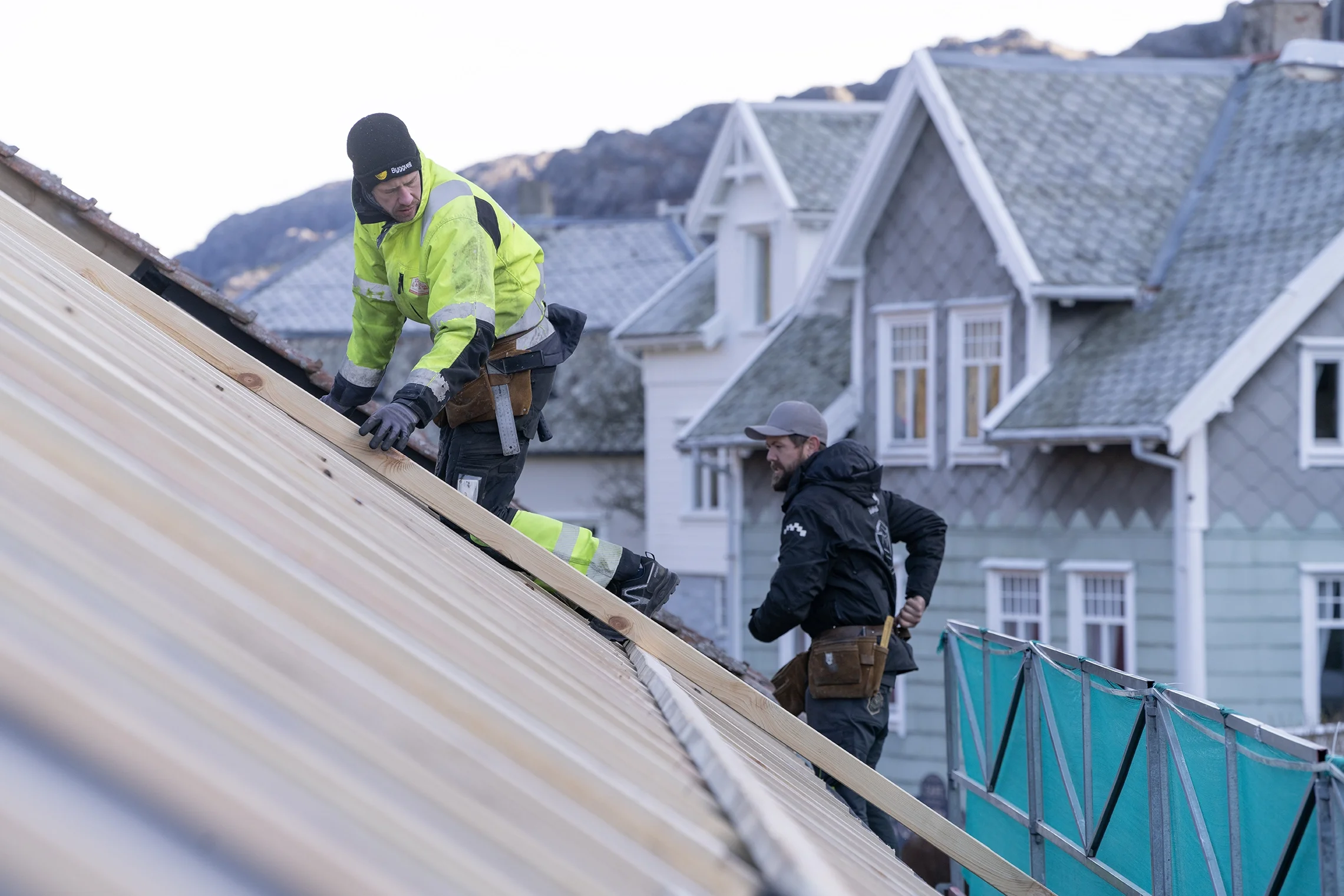 Two construction workers installing or repairing a wooden roof on a residential building with nearby houses and mountainous landscape in the background.