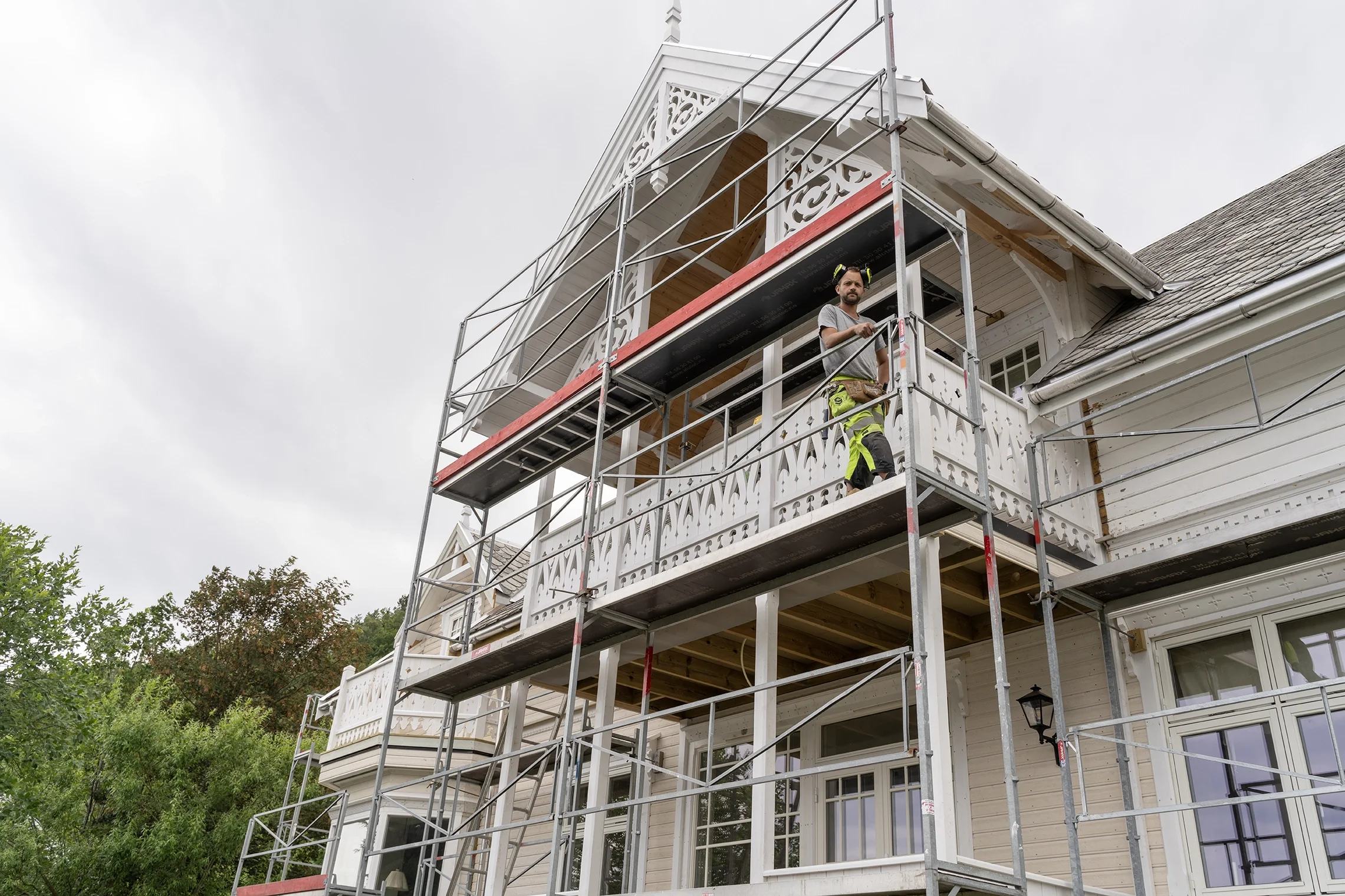 Construction workers installing or repairing the upper balcony of a house on scaffolding, with cloudy sky in the background.