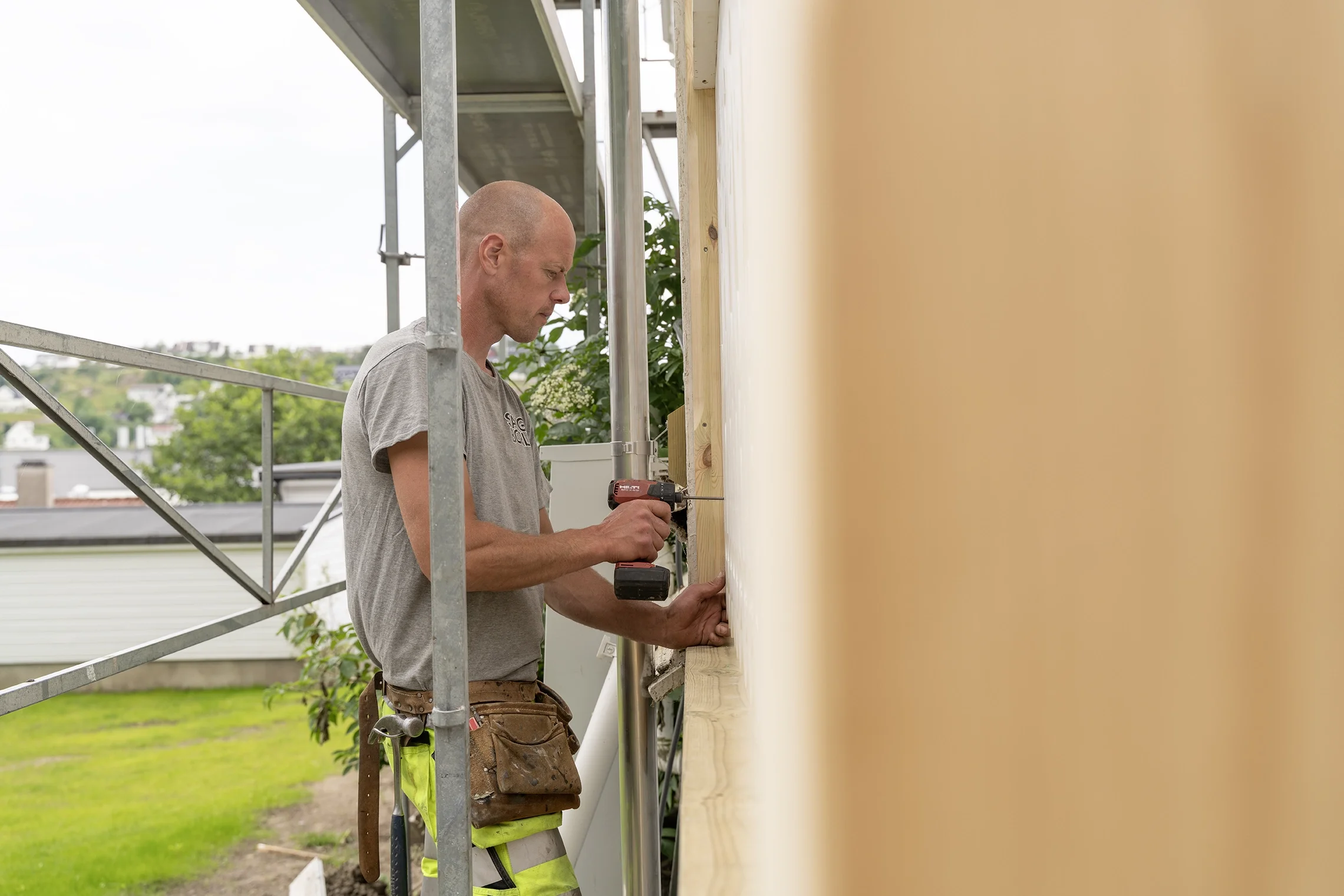 A man using a cordless drill on a wooden fence outside a house, with scaffolding structure nearby.