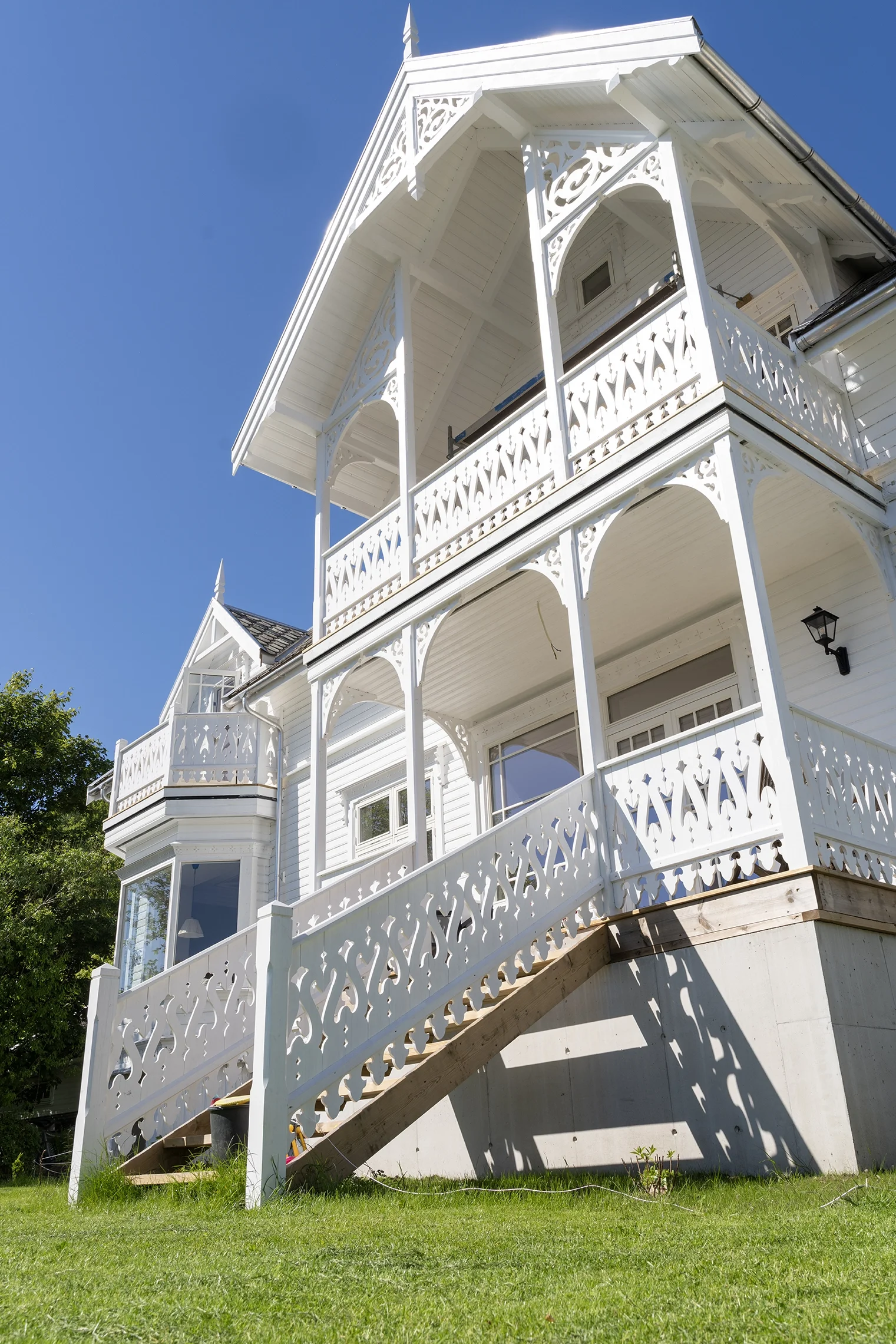 White Victorian-style house with ornate wooden balconies, decorative trim, and a front staircase. Clear blue sky and green lawn in foreground.