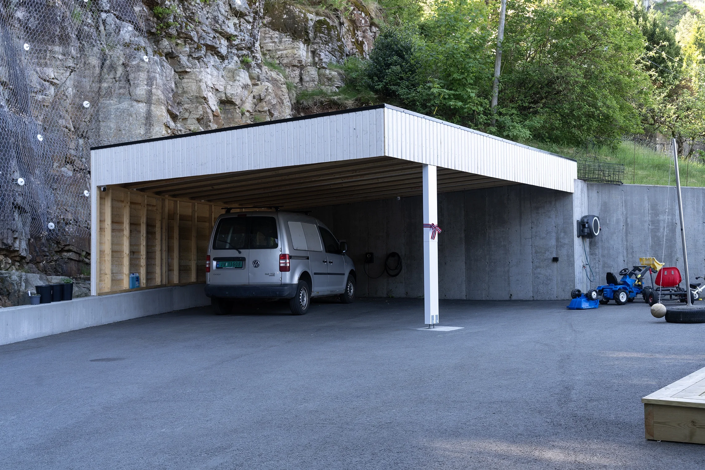 A modern carport with a gray Volkswagen van parked underneath. The structure has a white metal roof supported by a single white post, with a wooden wall on one side and a concrete wall on the other. To the right, there are children's toys, including