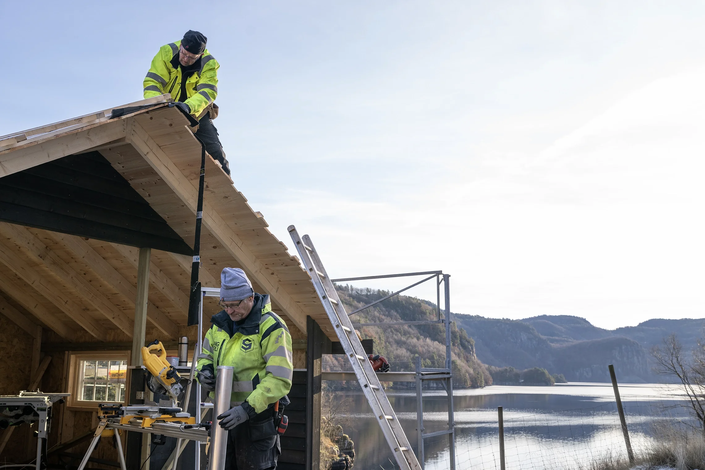 Two construction workers building a wooden structure outdoors near a lake with mountains in the background. One worker is on the roof, and the other is working with tools on the ground.